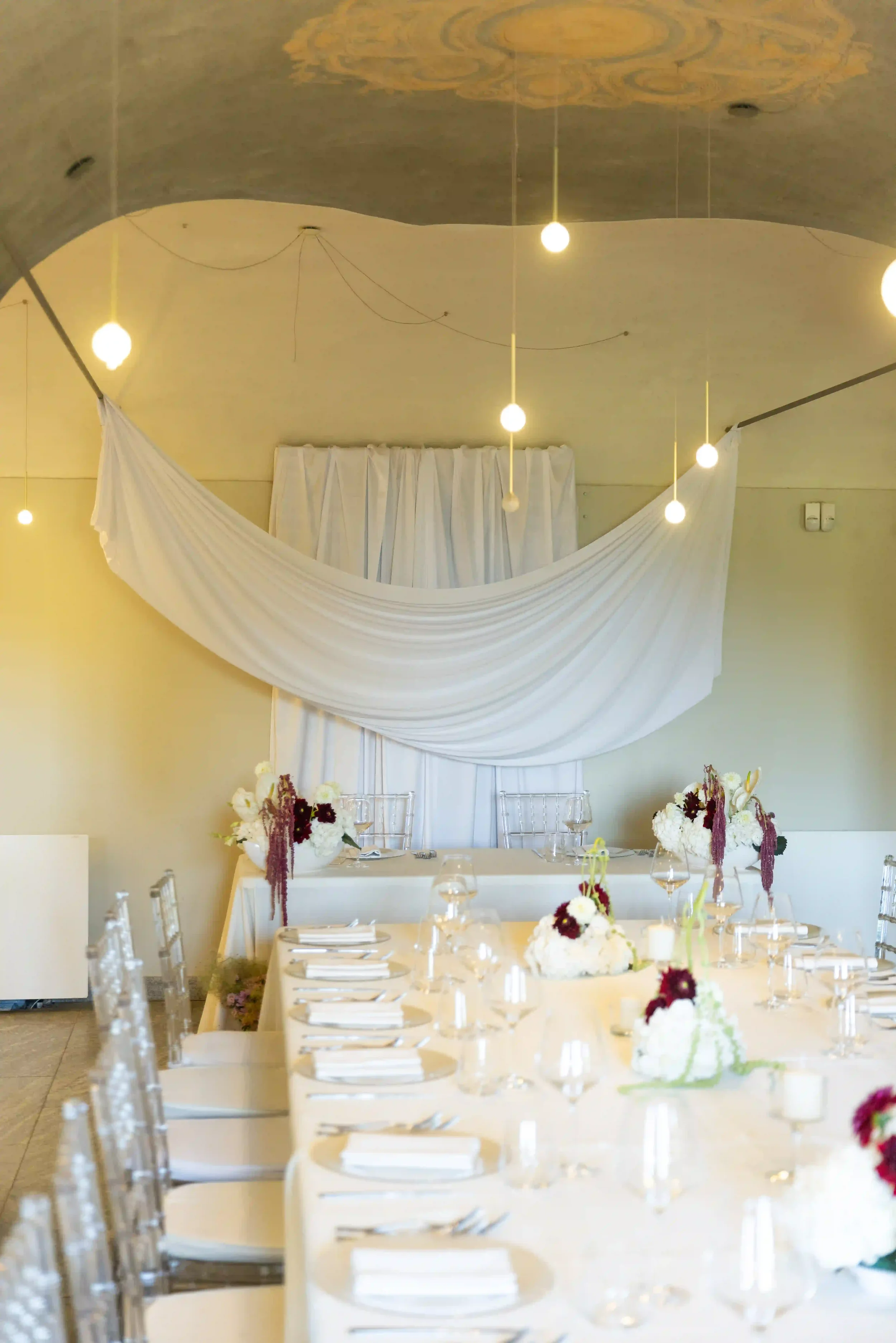 Allestimento della sala da pranzo della villa con uno scenografico drappeggio di tessuto bianco sulla parete di fondo, punti luce sospesi e tavoli imperiali decorati.