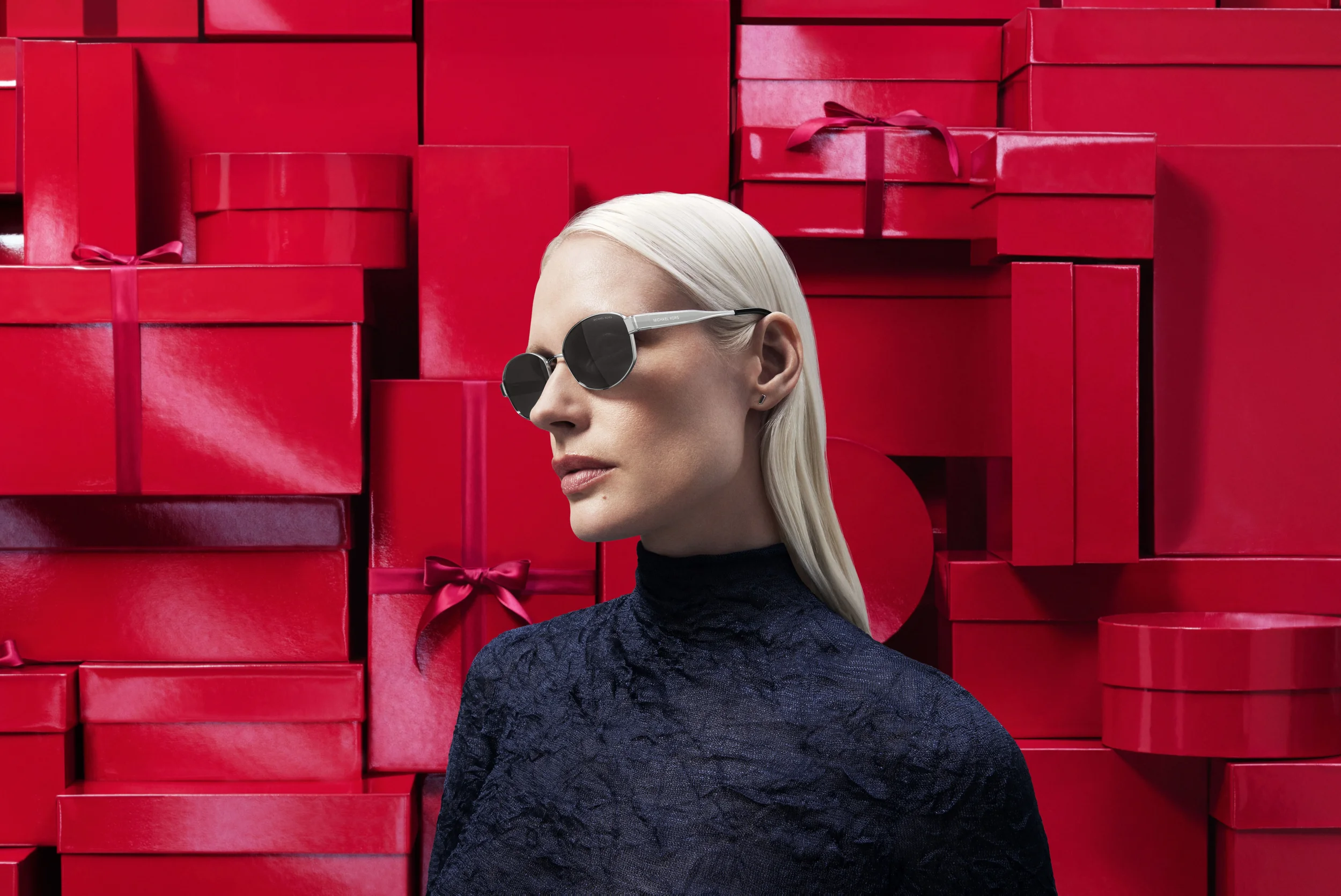 Ritratto di profilo della modella bionda che indossa occhiali da sole ovali scuri con dettagli metallici argentati, in posa davanti a una pareti di pacchi regalo rossi per Sunglass Hut.