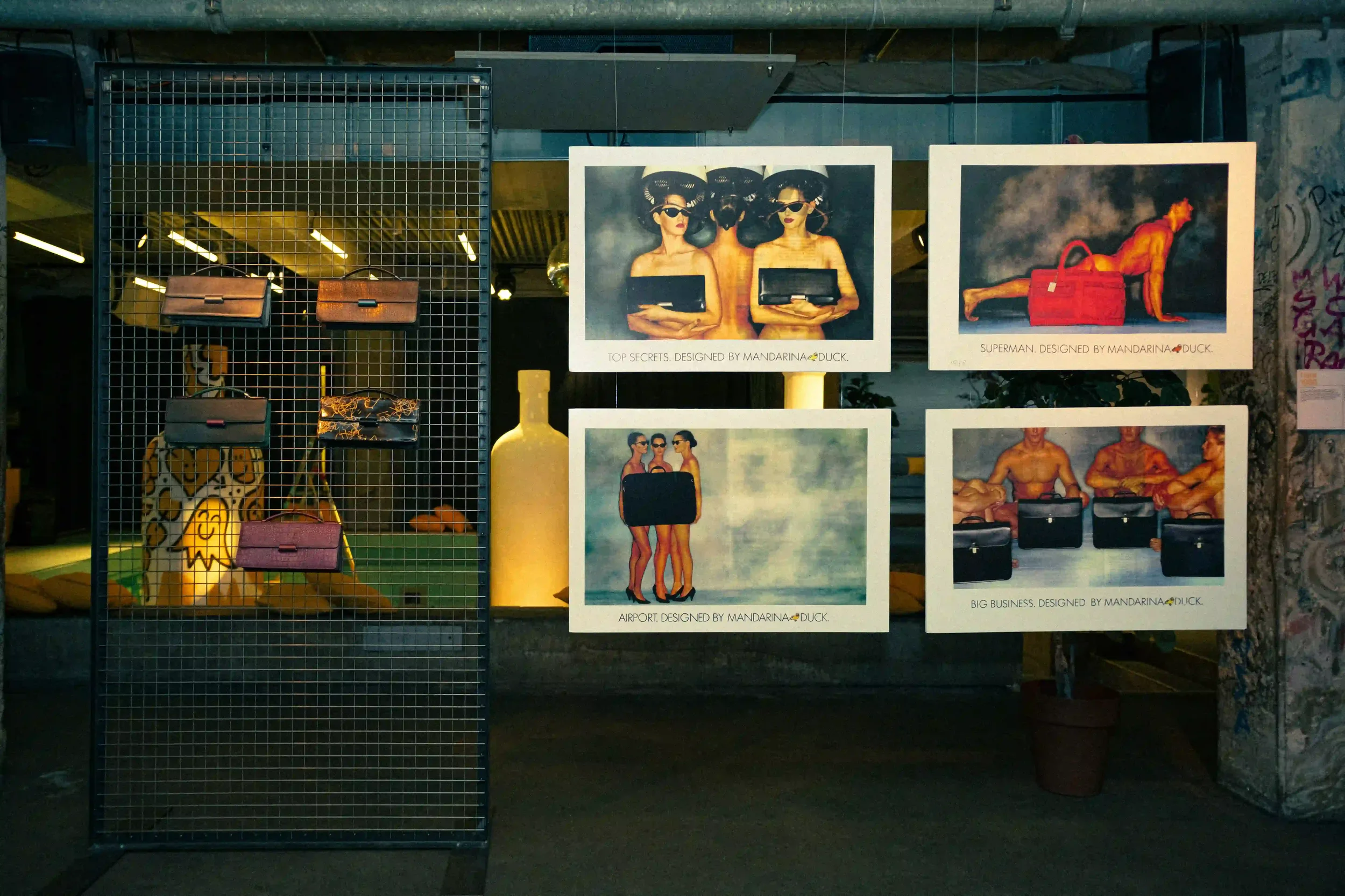 Allestimento scenografico per la mostra d'archivio Mandarina Duck a Berlino, con borse storiche esposte su griglia metallica e poster pubblicitari sospesi.