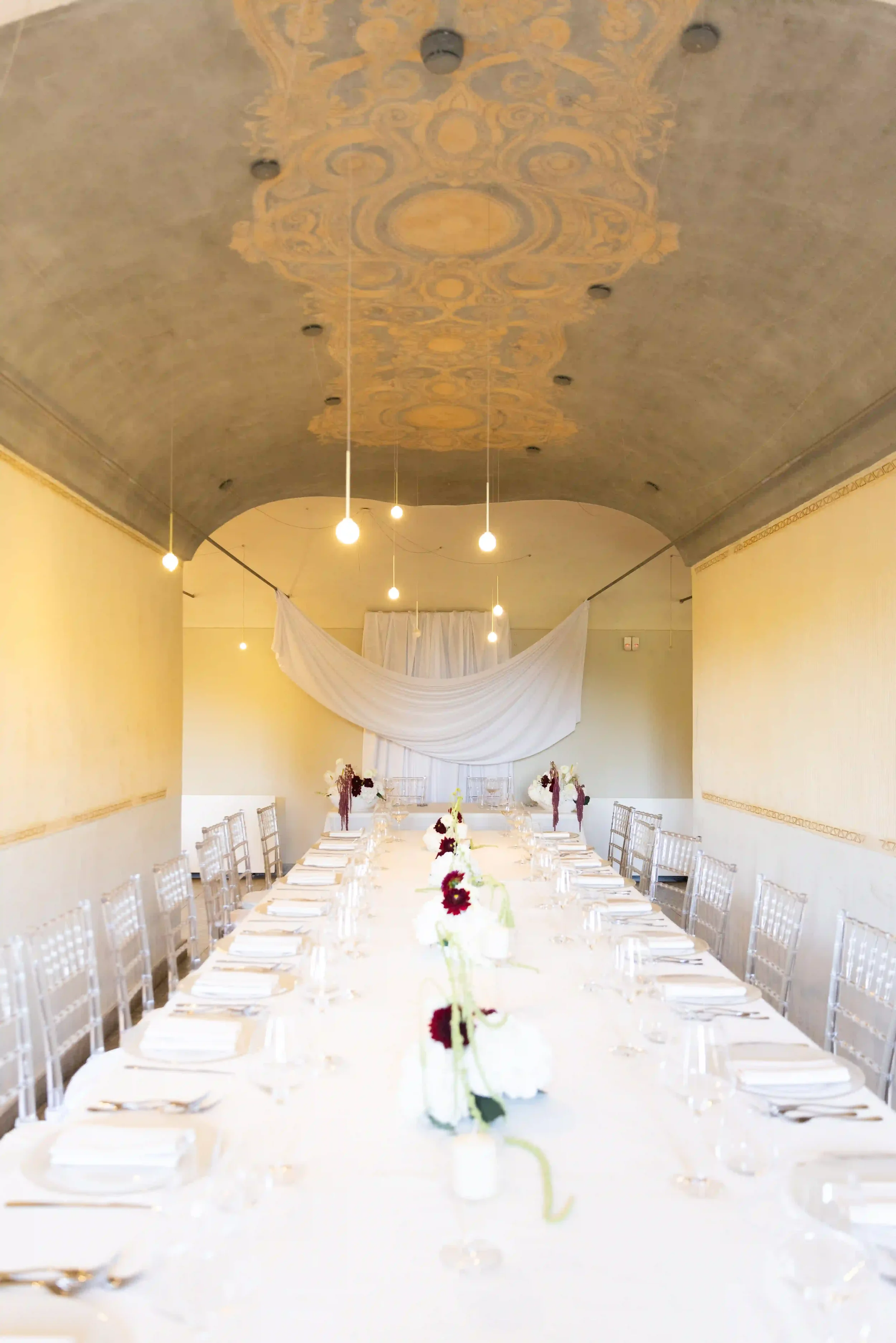 Inquadratura simmetrica della sala ricevimenti con soffitto decorato a volta, lungo tavolo imperiale apparecchiato in tono monocromatico bianco e sedie Ghost trasparenti.