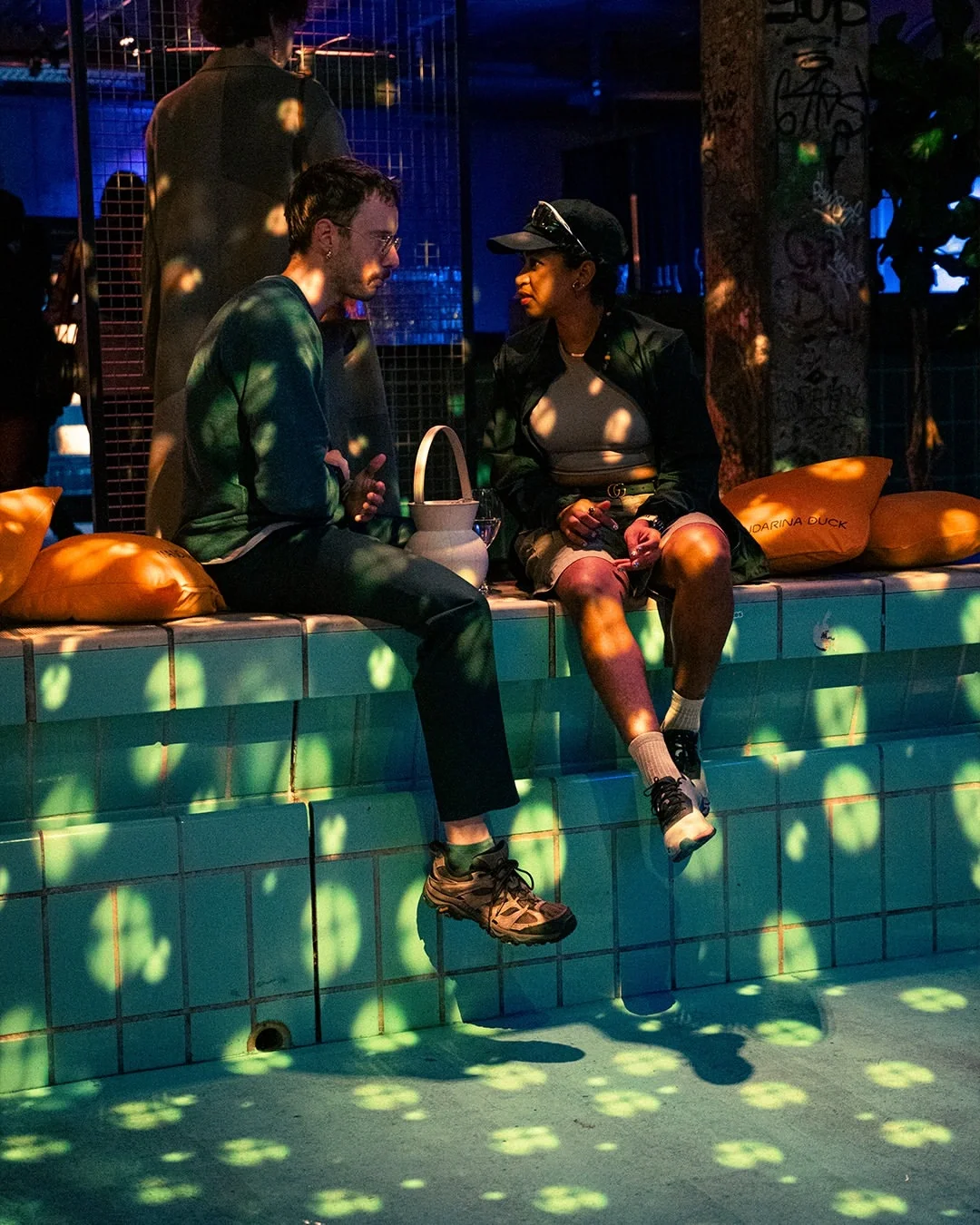 Area lounge scenografica per l'evento Mandarina Duck a Berlino, con cuscini brandizzati arancioni posizionati lungo il bordo di una struttura azzurra in piastrelle.