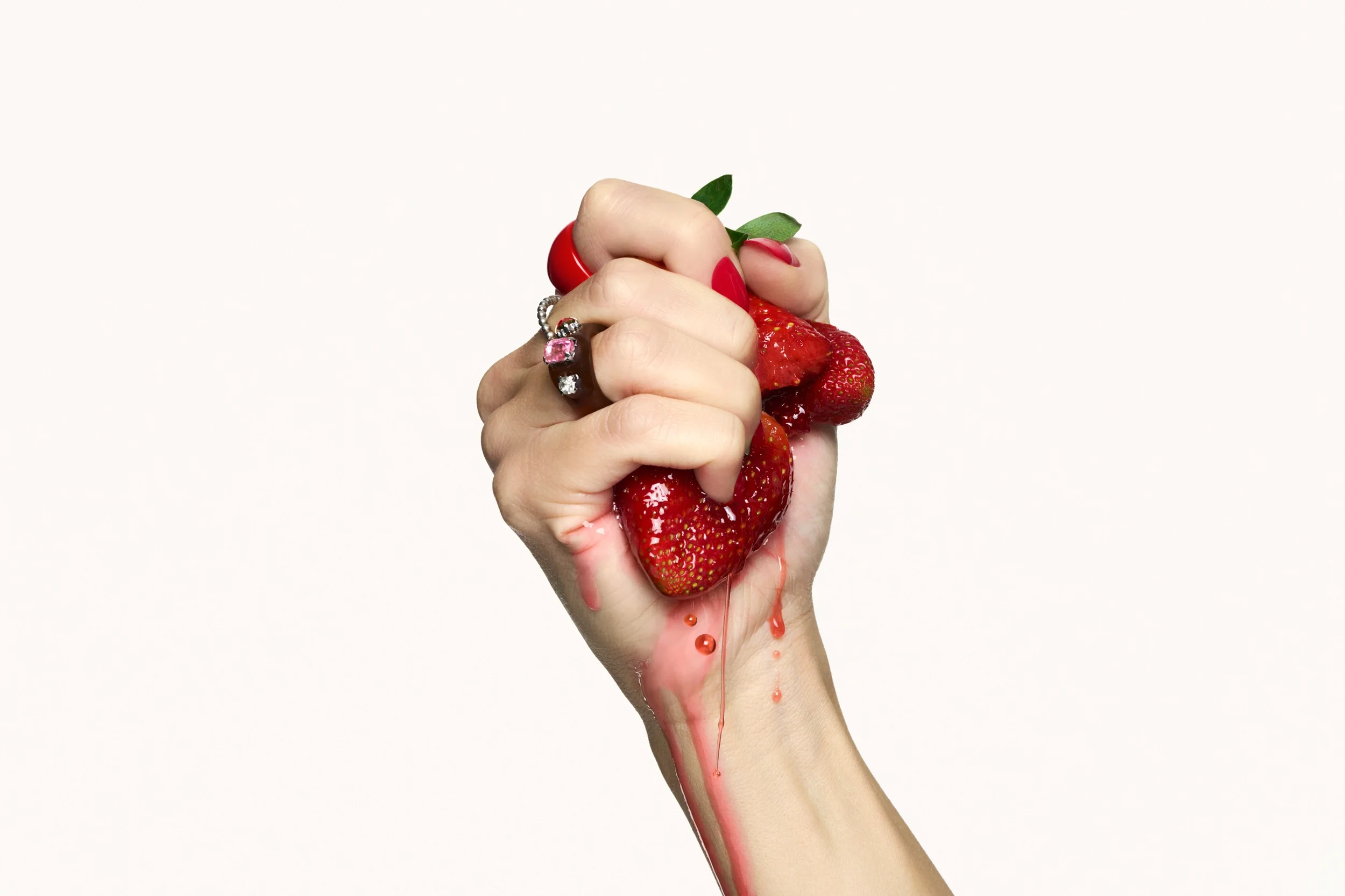Scatto still life d'impatto per la campagna beauty Pupa Milano con mano impreziosita da anelli che schiaccia una fragola fresca lasciando colare il succo lungo il polso su sfondo bianco.