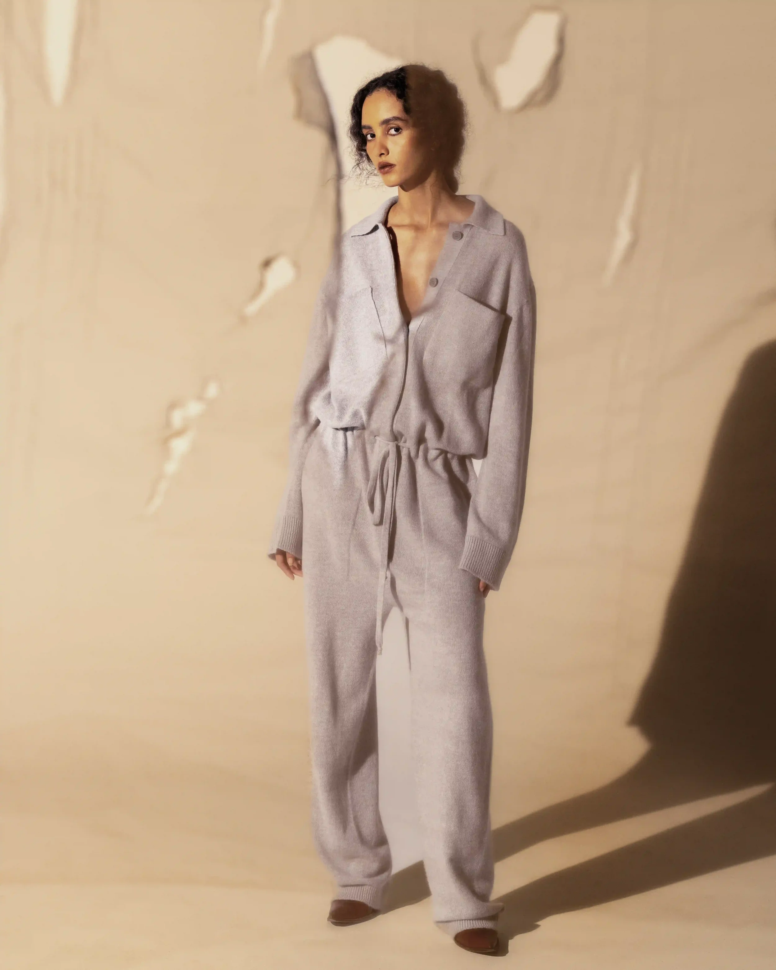 Scenografia e props styling per editorial moda Laneus: modella con jumpsuit morbida in lana grigio chiaro adagiata su un fondale materico con intagli e ombre radenti.