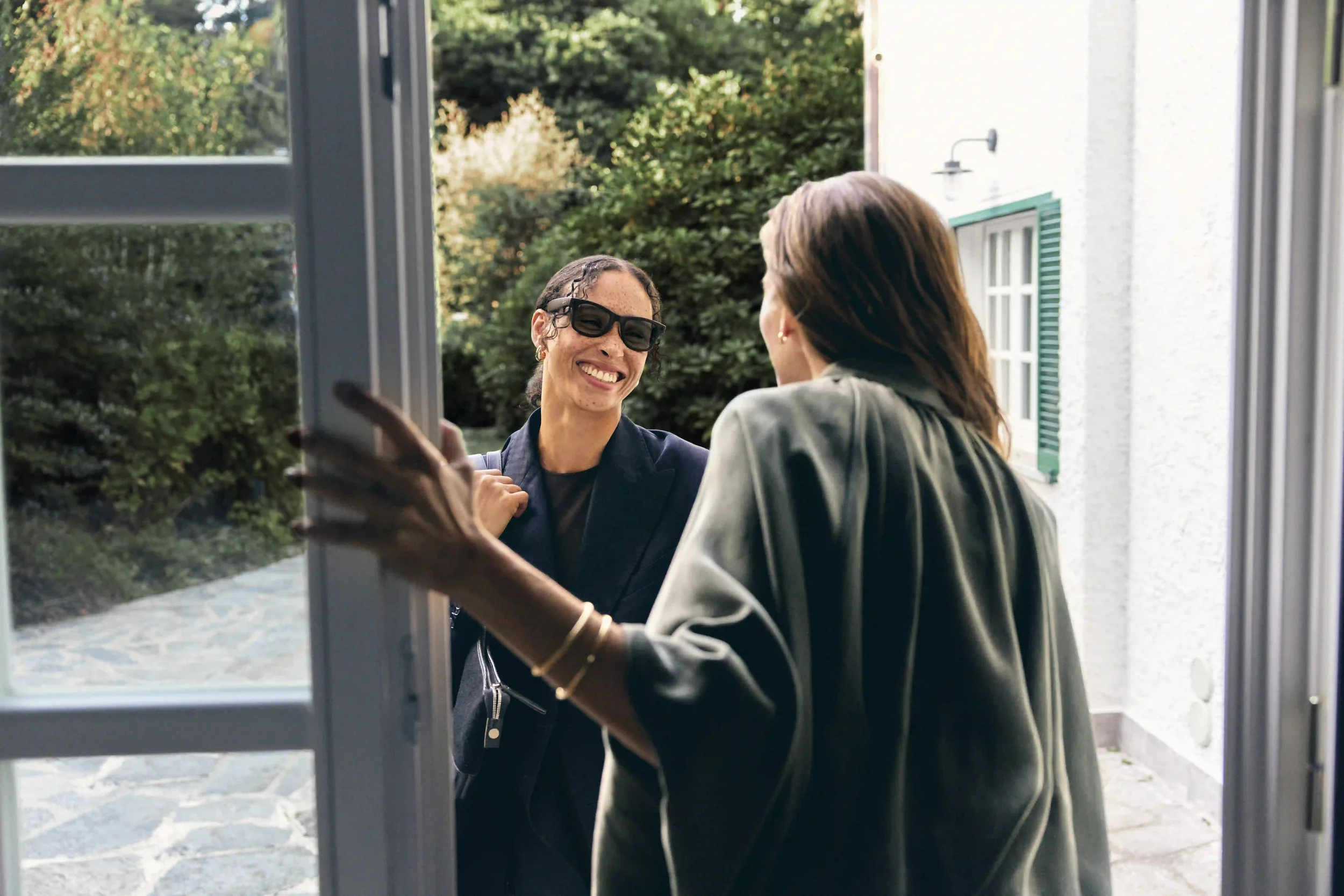 Fotografia lifestyle e promozionale per la campagna natalizia Sunglass Hut con modella che accoglie un'amica aprendo la porta di casa, con ospite sorridente che indossa occhiali da sole neri di tendenza.