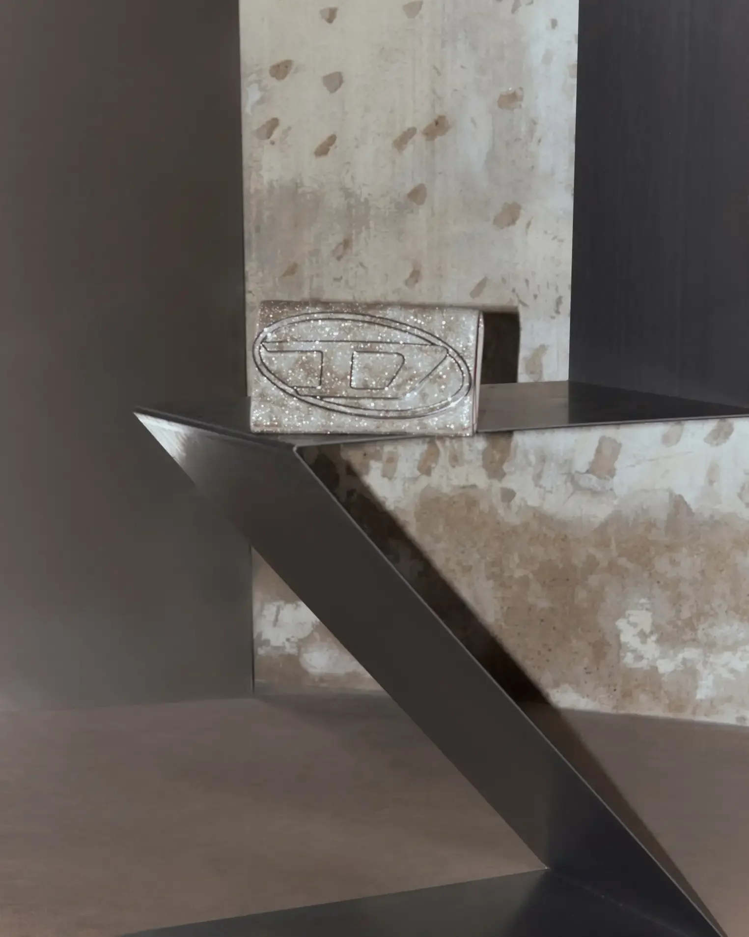 Fotografia still life di una borsa pochette Diesel ricoperta di glitter argento con l'iconico logo metallico a rilievo, adagiata su una struttura geometrica in acciaio scuro con sfondo di cemento d'archivio. Set design per Le Mile Magazine.