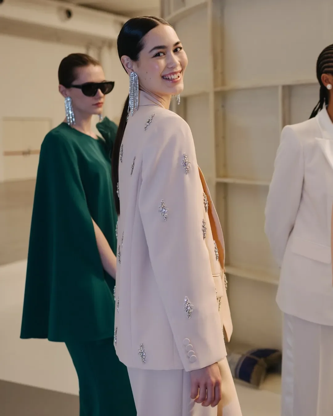 Ritratto backstage di modella sorridente in giacca sartoriale rosa cipria impreziosita da ricami in cristallo e orecchini pendenti scintillanti, durante i preparativi della sfilata Marina Rinaldi.