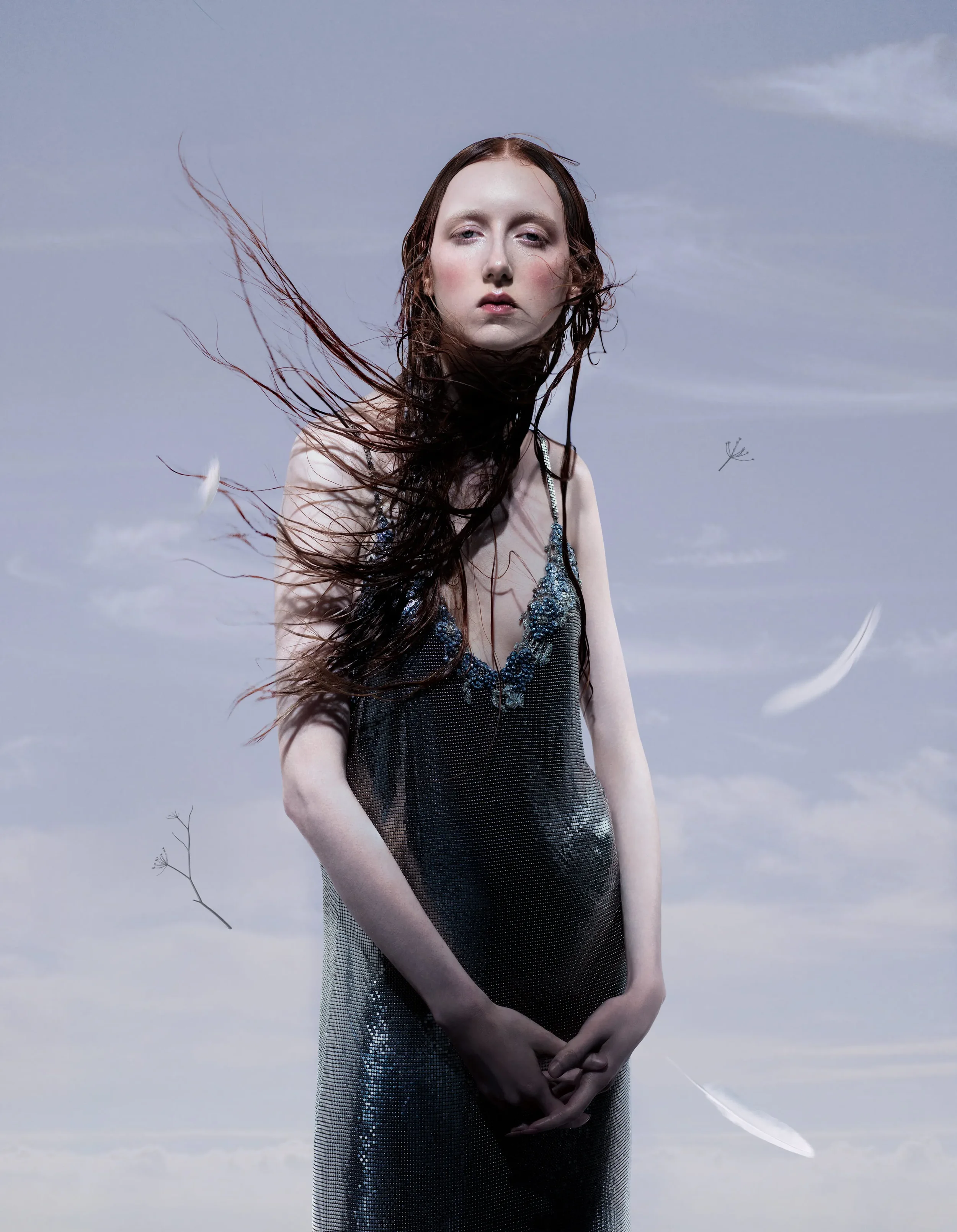 Primo piano editoriale per Vogue Portugal di una modella con capelli mossi dal vento che indossa un abito scivolato in maglia metallica con cristalli, tra piume ed elementi botanici fluttuanti.