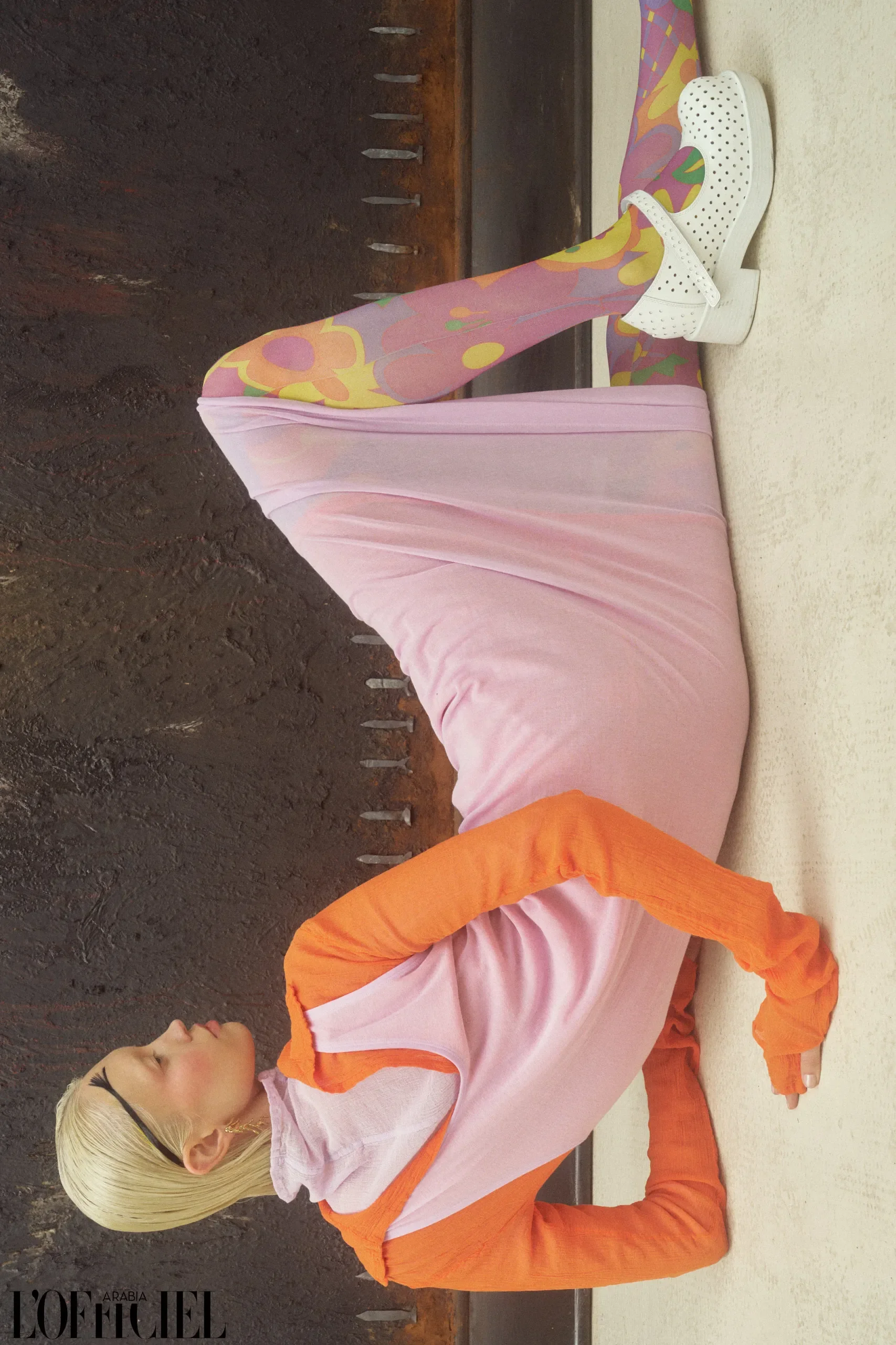 Posa acrobatica della modella stesa a terra con abito rosa, manicotti arancioni, collant floreali multicolor e scarpe bianche traforate con zeppa."