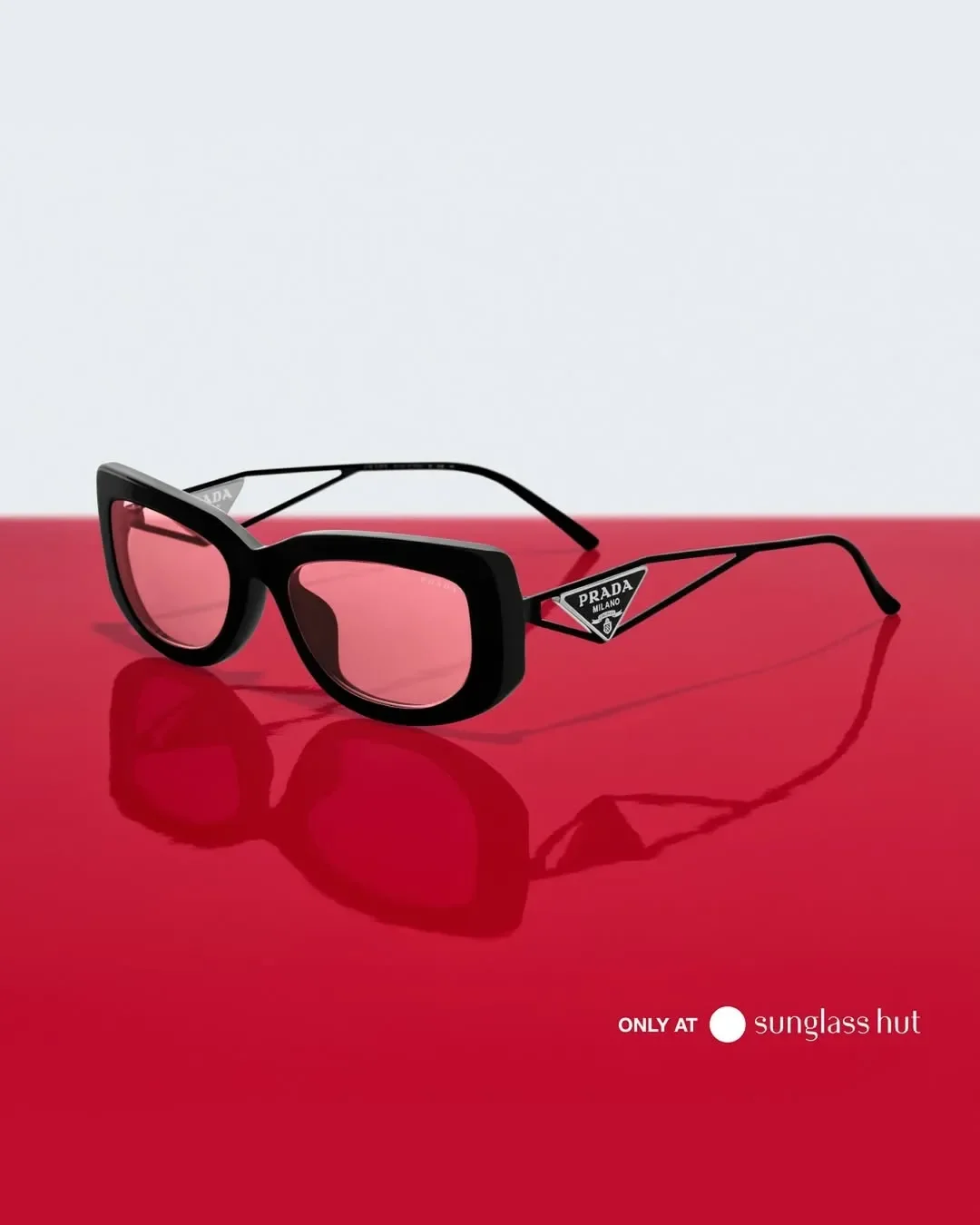 Scatto still life con occhiali da sole Prada cat-eye neri con lenti rosa e logo triangolare sull'asta, adagiati su una superficie rossa riflettente per la campagna Sunglass Hut.