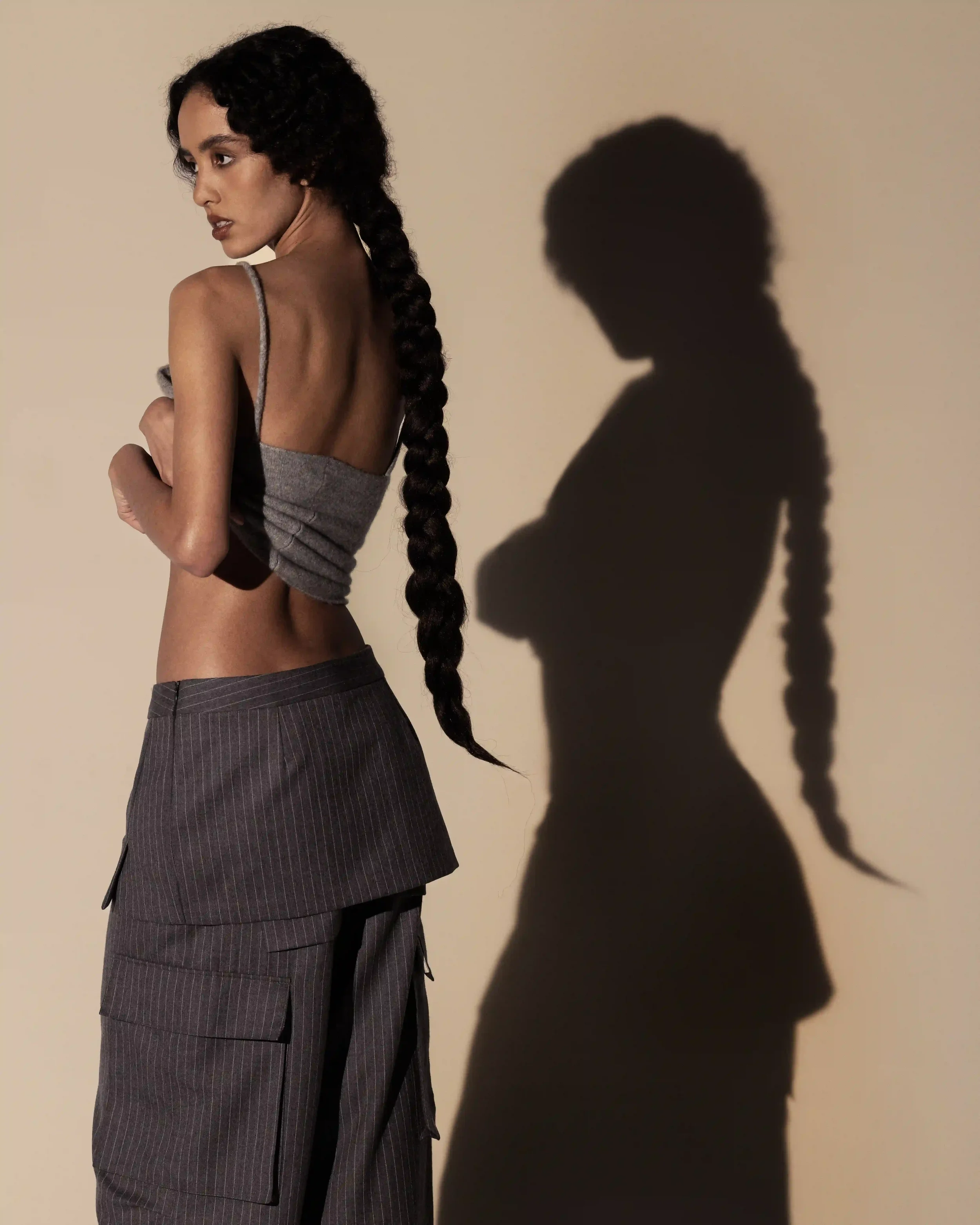 "Set design minimale con ombra netta e marcata per la campagna Laneus. Modella ritratta di spalle con top in lana grigia, pantaloni gessati e treccia scenografica