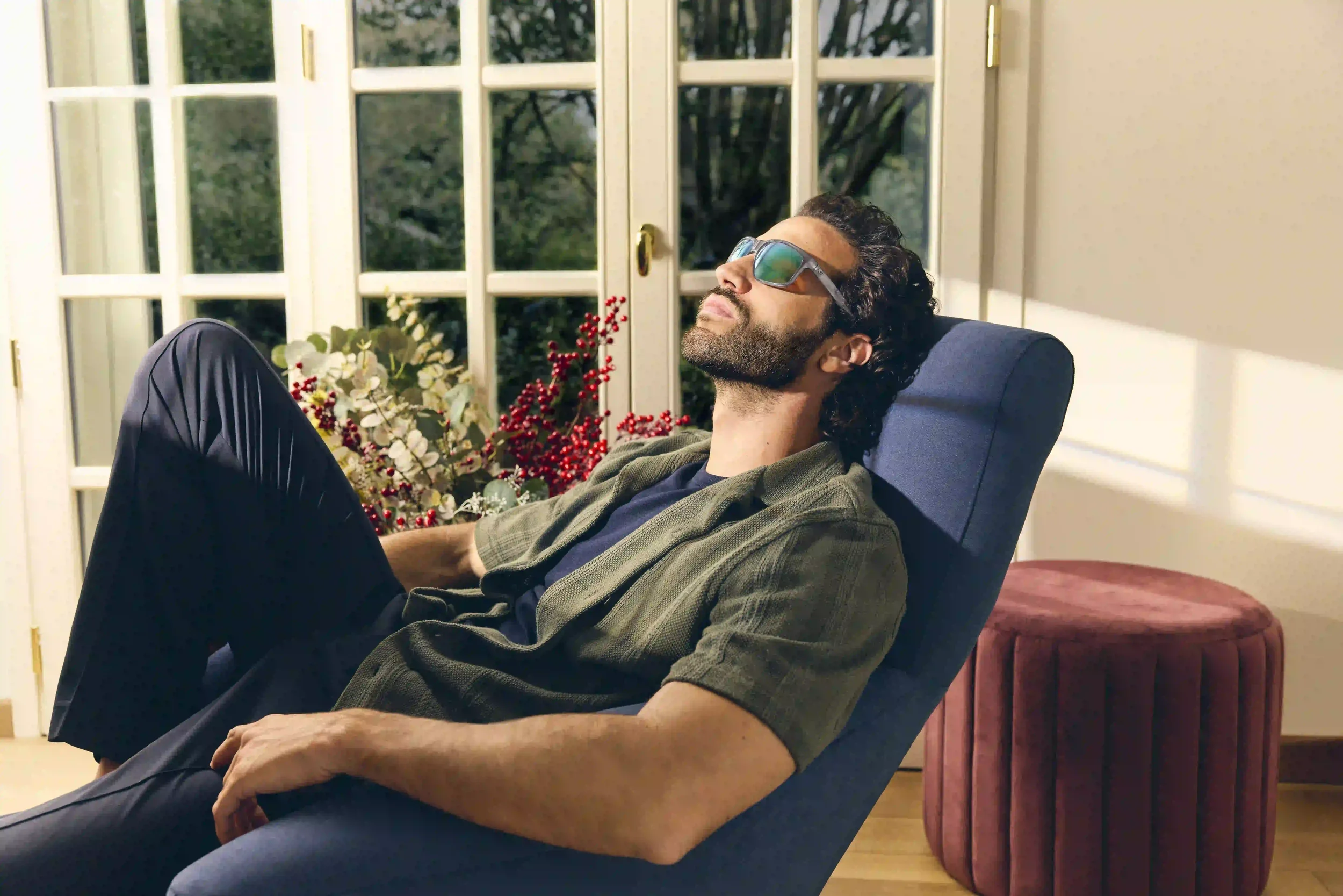 Ritratto lifestyle per la campagna natalizia Sunglass Hut con modello rilassato su chaise longue blu accanto a una finestra e composizione di bacche rosse, con occhiali da sole sportivi dalle lenti a specchio.