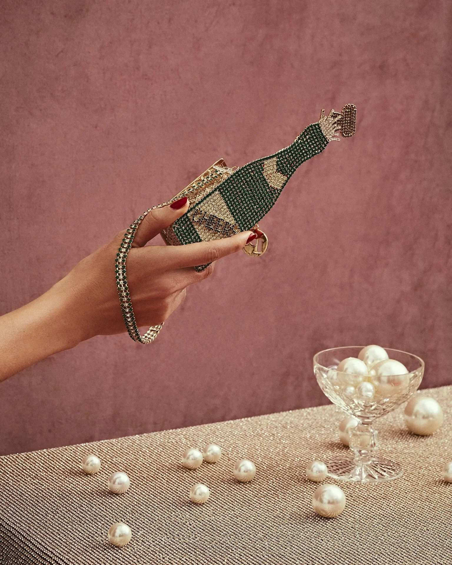 Fotografia still life della campagna Rosantica con mano che sostiene una borsa gioiello a forma di bottiglia di champagne tra perle sparse e una coppa di cristallo.