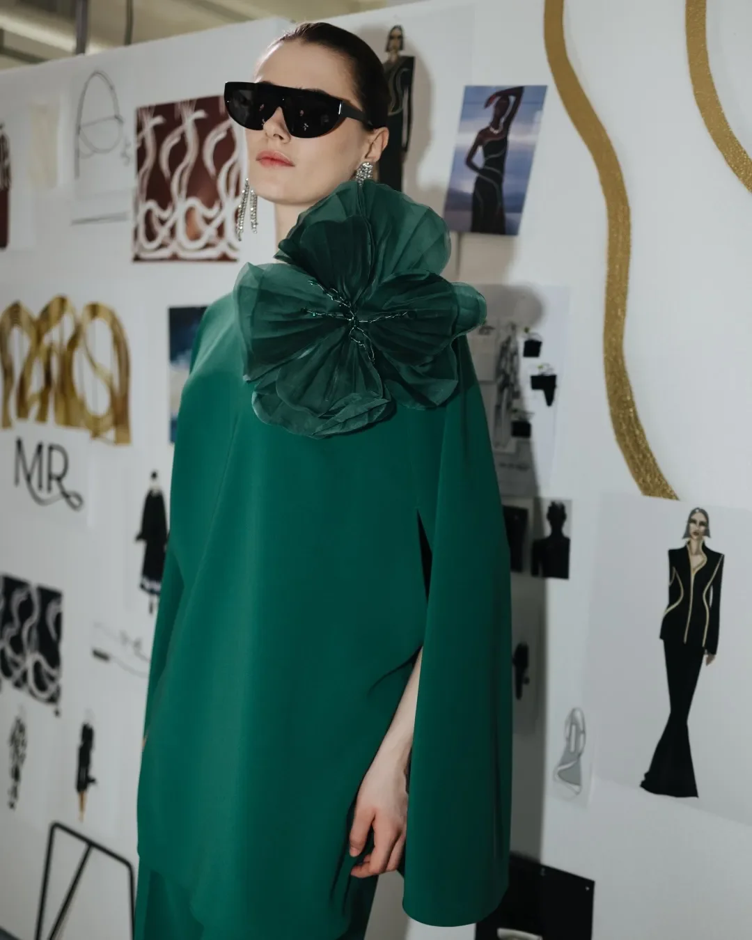 Modella nel backstage della sfilata Marina Rinaldi indossa un abito stile cappa verde smeraldo con grande applicazione floreale e occhiali da sole, posando davanti al pannello espositivo con bozzetti e studi di collezione