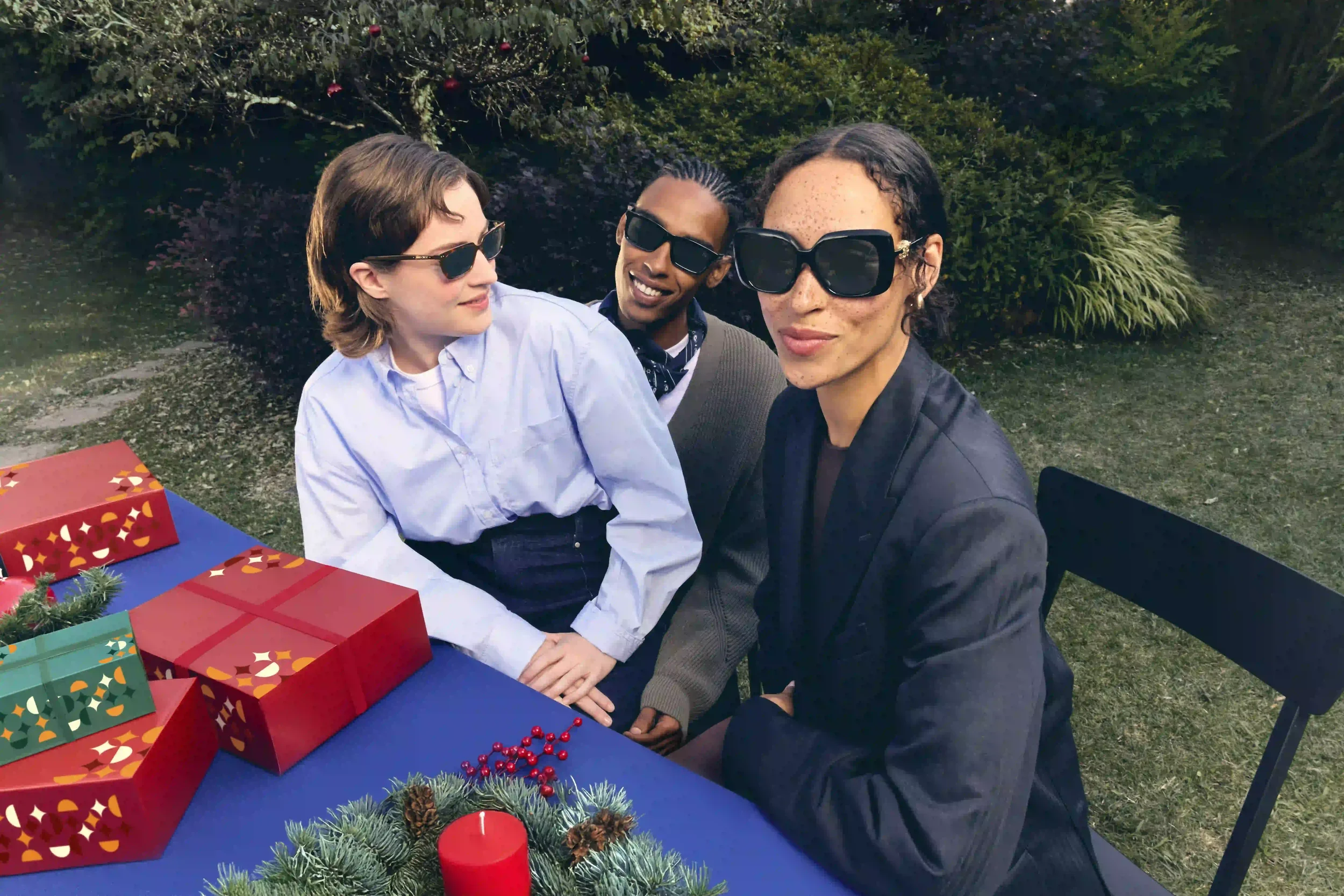 Fotografia promozionale Sunglass Hut con tre ragazzi seduti al tavolo da giardino allestito per il Natale con ghirlanda e pacchi regalo colorati, tutti indossando occhiali da sole oversize e classici.