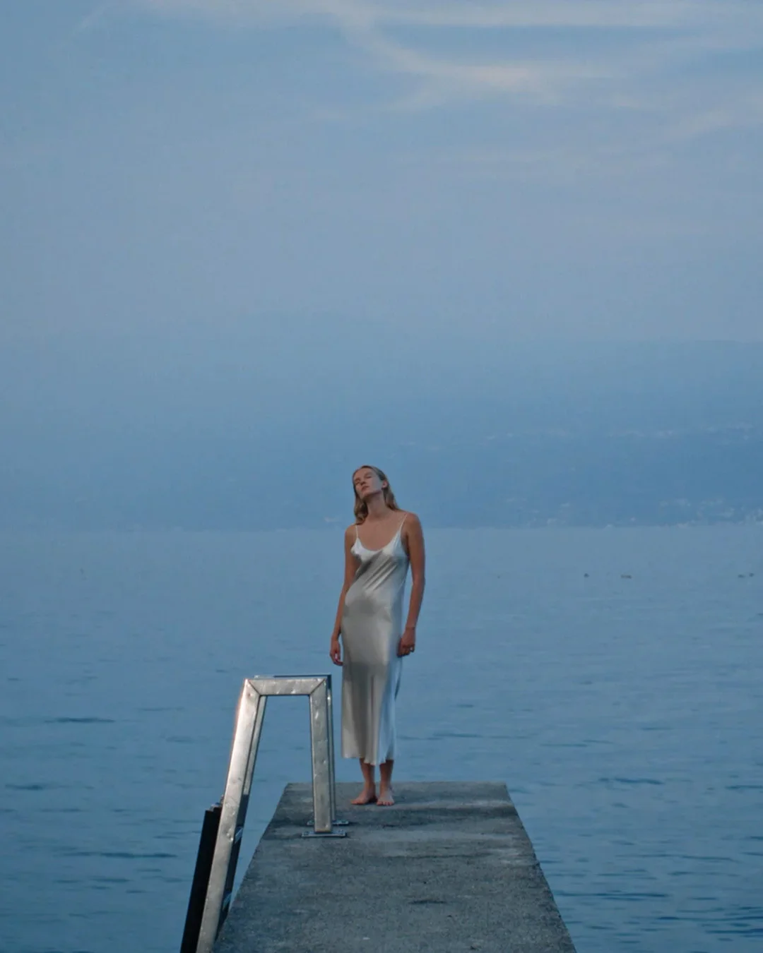 Fotografia editoriale di moda con modella in abito scivolato in seta tonalità argento, posizionata all'estremità di un molo in cemento al tramonto sul lago.