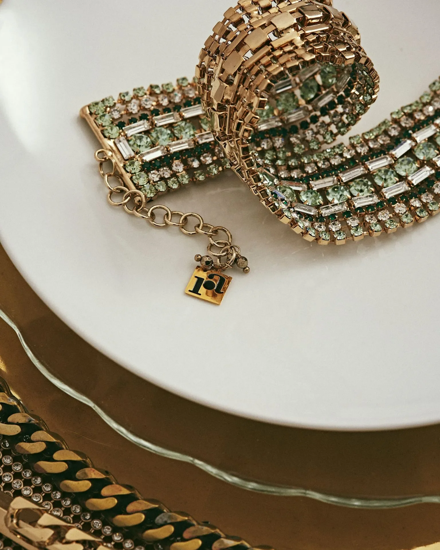 Dettaglio still life per la campagna Rosantica con bracciale gioiello con cristalli verdi e charm logato adagiato su un piatto bianco.
