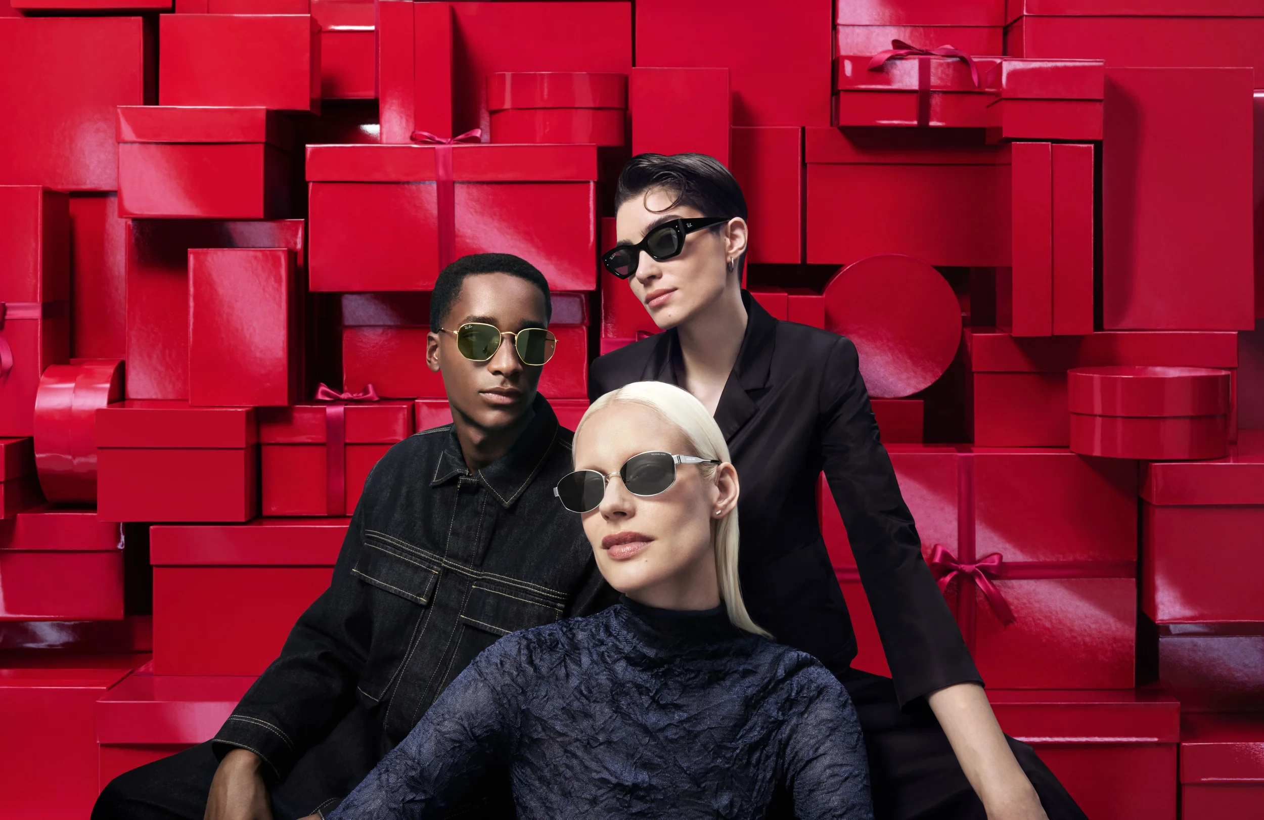 Scatto di campagna editoriale per Sunglass Hut con tre modelli in posa che indossano occhiali da sole di lusso, davanti a un imponente sfondo scenografico composto da scatole regalo rosse natalizie.