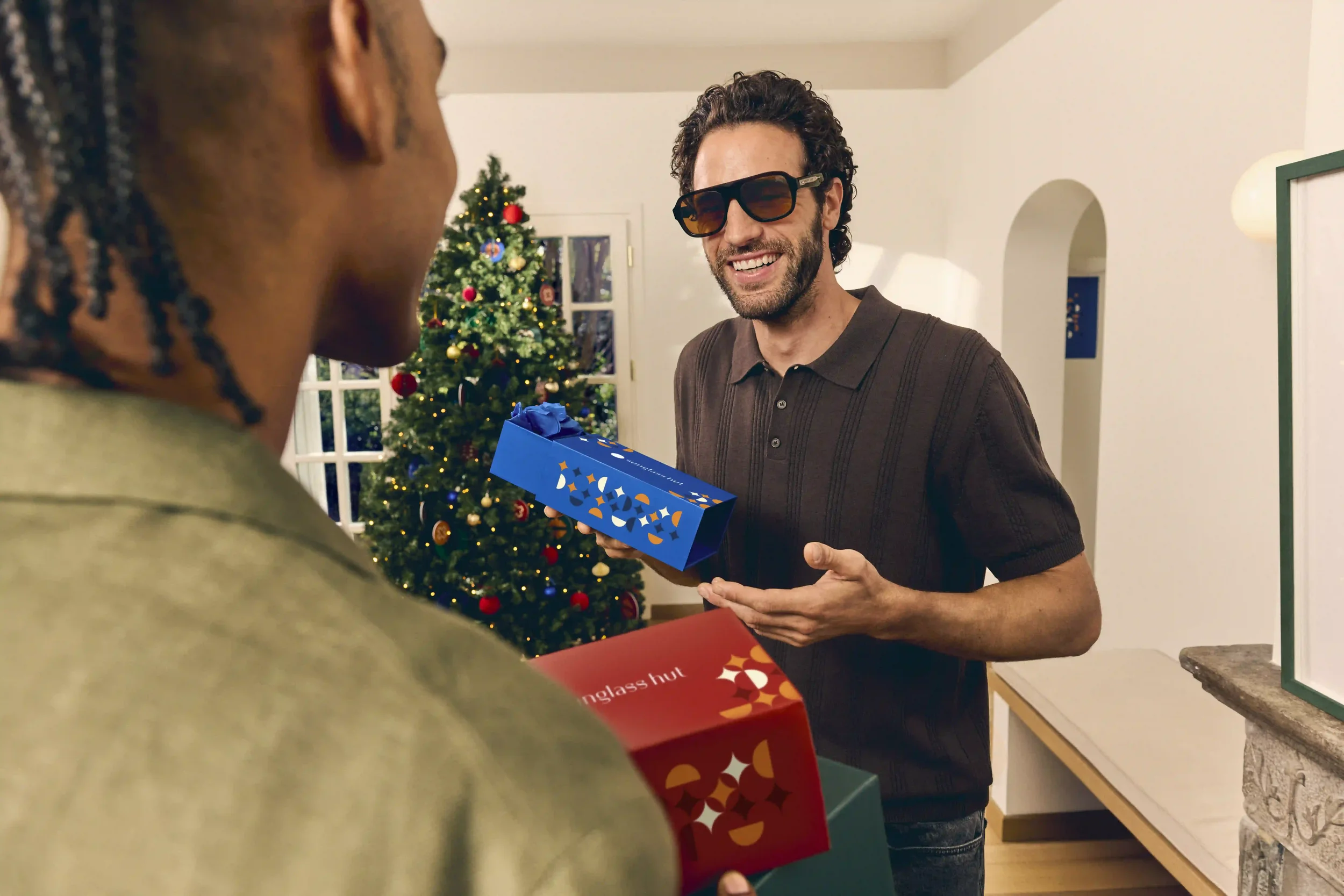 Scatto promozionale natalizio Sunglass Hut con scambio di scatole regalo colorate tra modelli, con occhiali da sole squadrati da uomo e albero di Natale festivo sullo sfondo.