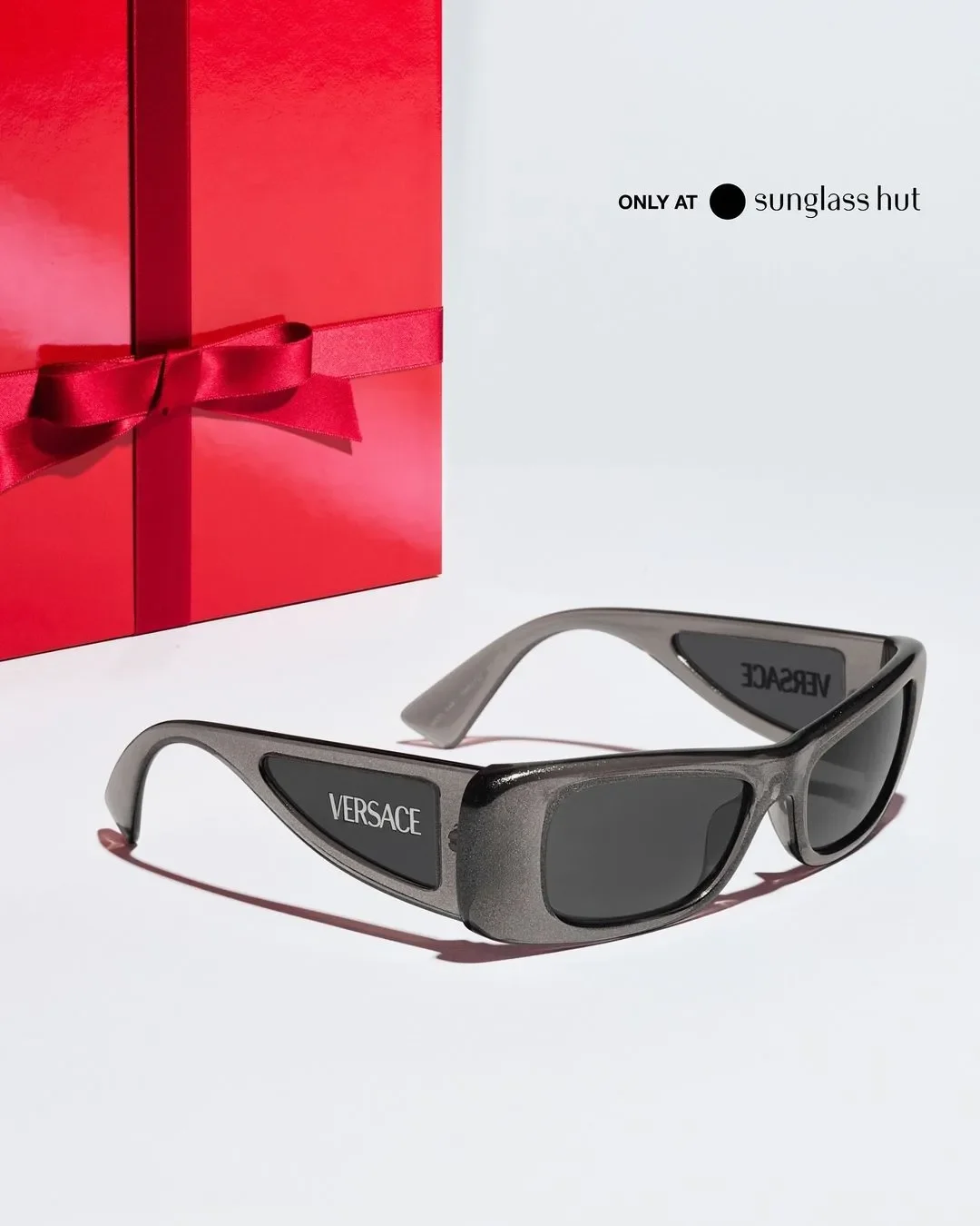 Inquadratura prospettica di occhiali da sole rettangolari Versace in finitura grigio metallizzato con logo laterale, disposti su piano bianco accanto a una scatola regalo rossa natalizia.