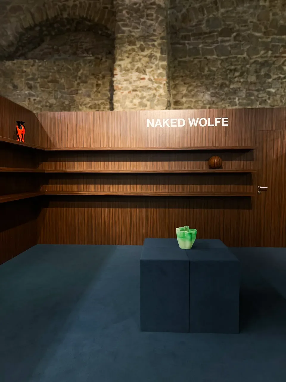 Allestimento pop-up retro-minimalista per Naked Wolfe con scaffalature in legno scuro, moquette blu, blocco espositivo centrale con vaso in vetro verde e logo a parete sotto architetture in mattoni a vista.