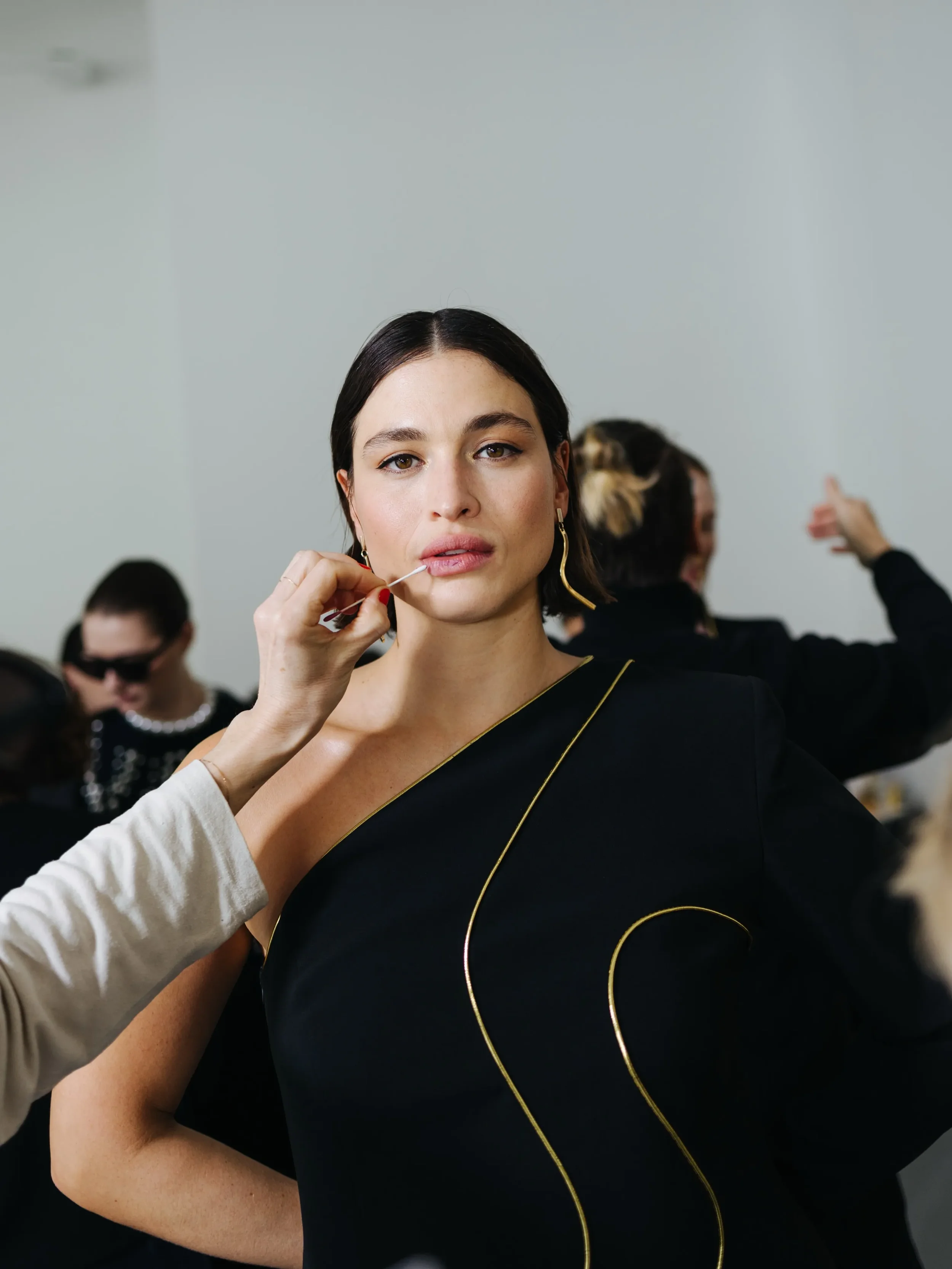 Momento di retropalco e beauty nel backstage Marina Rinaldi: la makeup artist applica il rossetto alla modella che indossa l'abito nero monospalla con dettagli oro