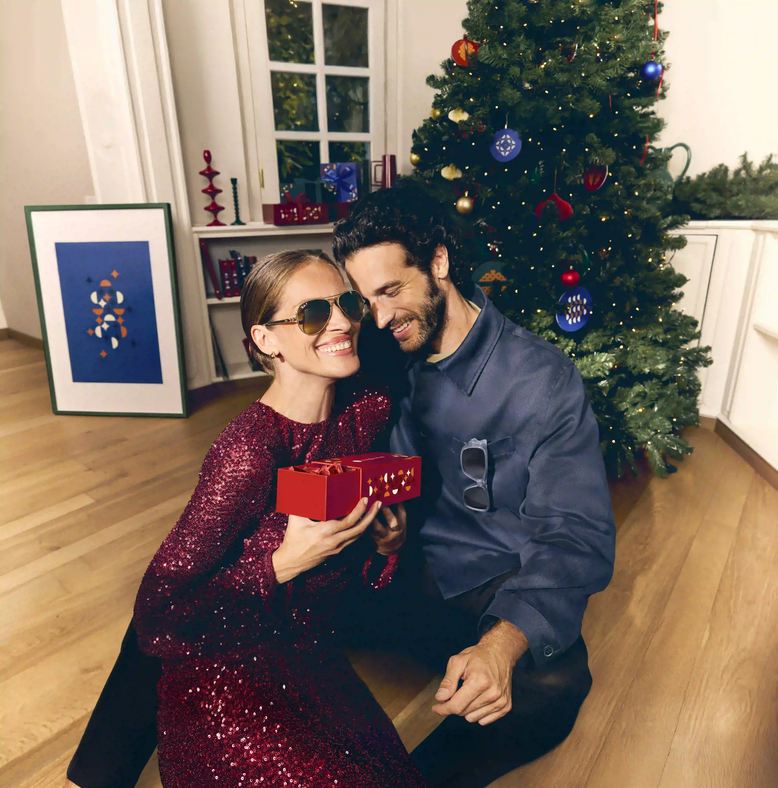 Fotografia di moda e party natalizio Sunglass Hut con modella in abito rosso di paillettes che indossa occhiali da sole a goccia dorati insieme al partner seduti vicino all'albero.