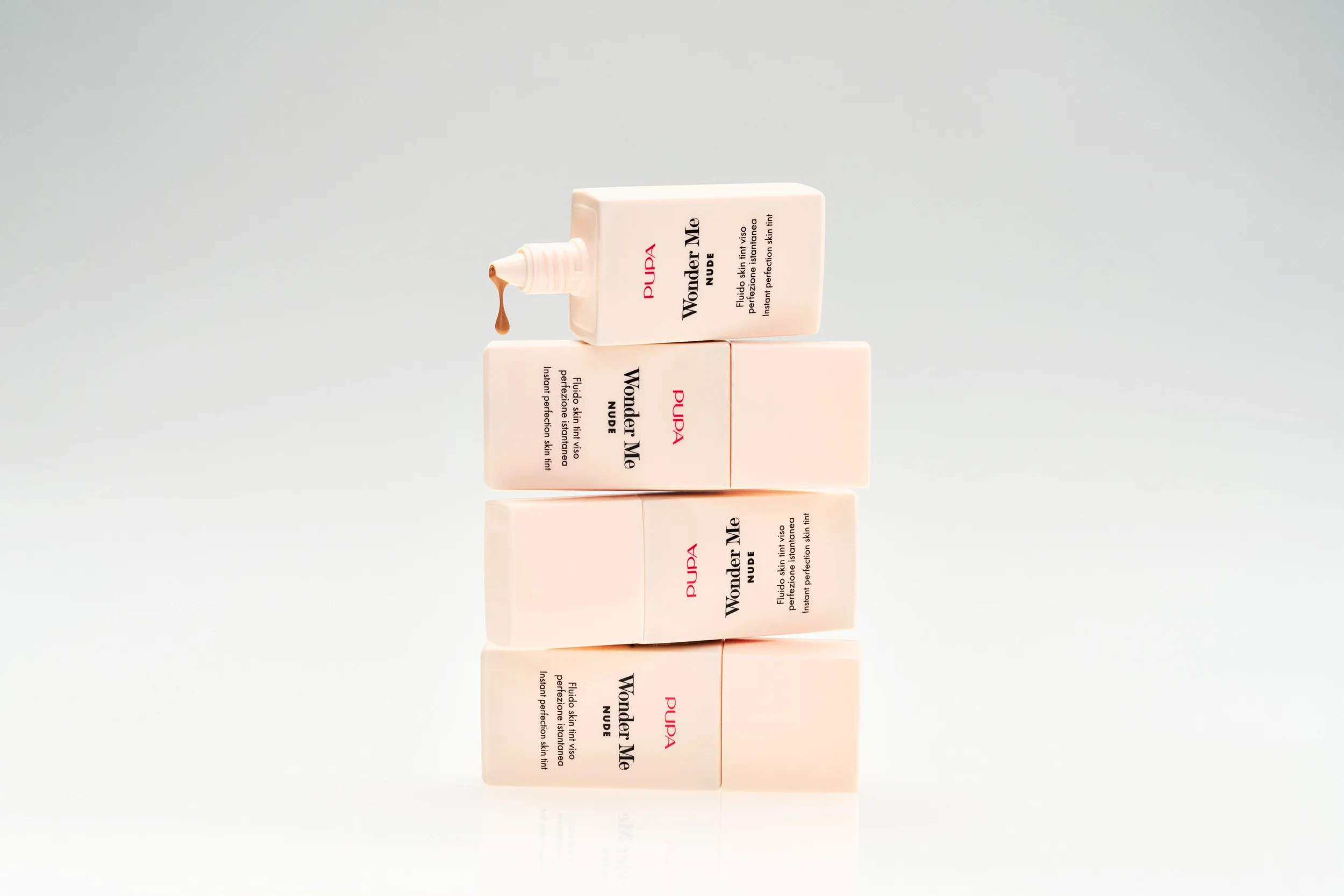 Scatto still life concettuale con quattro flaconi di Pupa Wonder Me Nude Skin Tint impilati in equilibrio verticale, con il flacone superiore aperto da cui fa capolino una goccia di fluido.