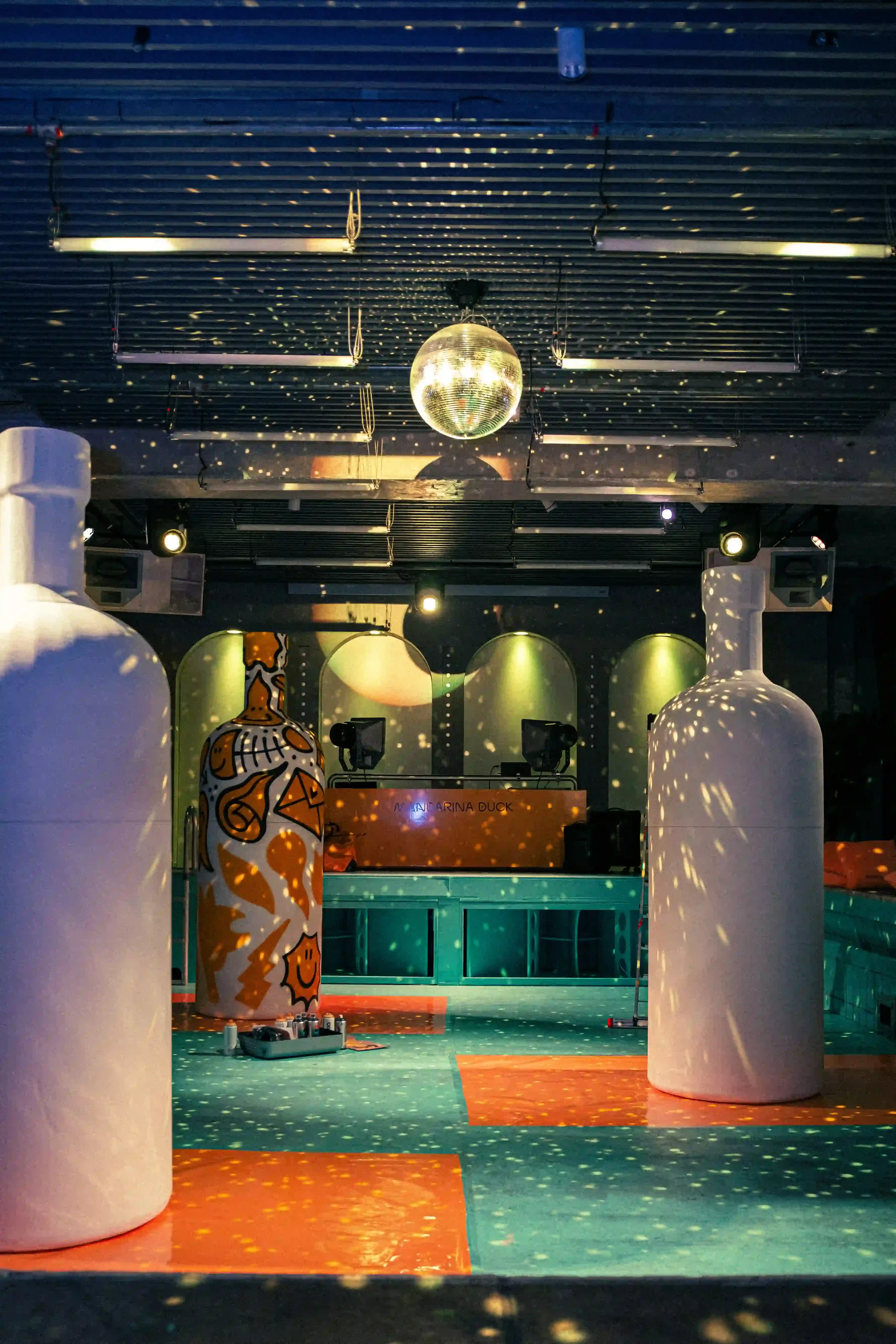 Set design per l'evento Mandarina Duck a Berlino con grandi sculture decorate, console DJ e strobosfera sospesa che riflette giochi di luce sul pavimento della location."