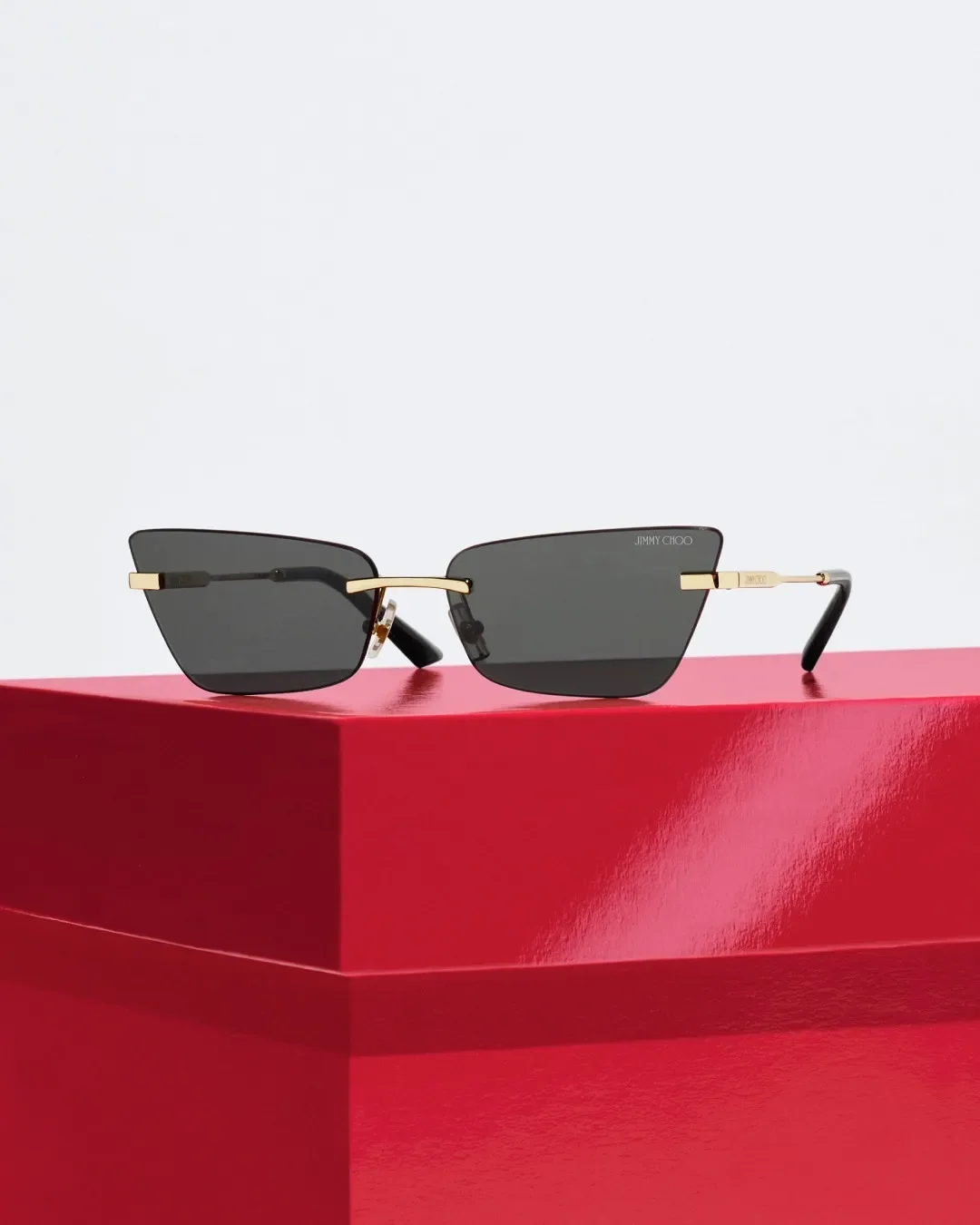 Scatto still life d'impatto con occhiali da sole cat-eye Jimmy Choo a giorno con dettagli dorati e lenti scure squadrate, adagiati su una scatola regalo lucida rossa per la campagna Sunglass Hut.
