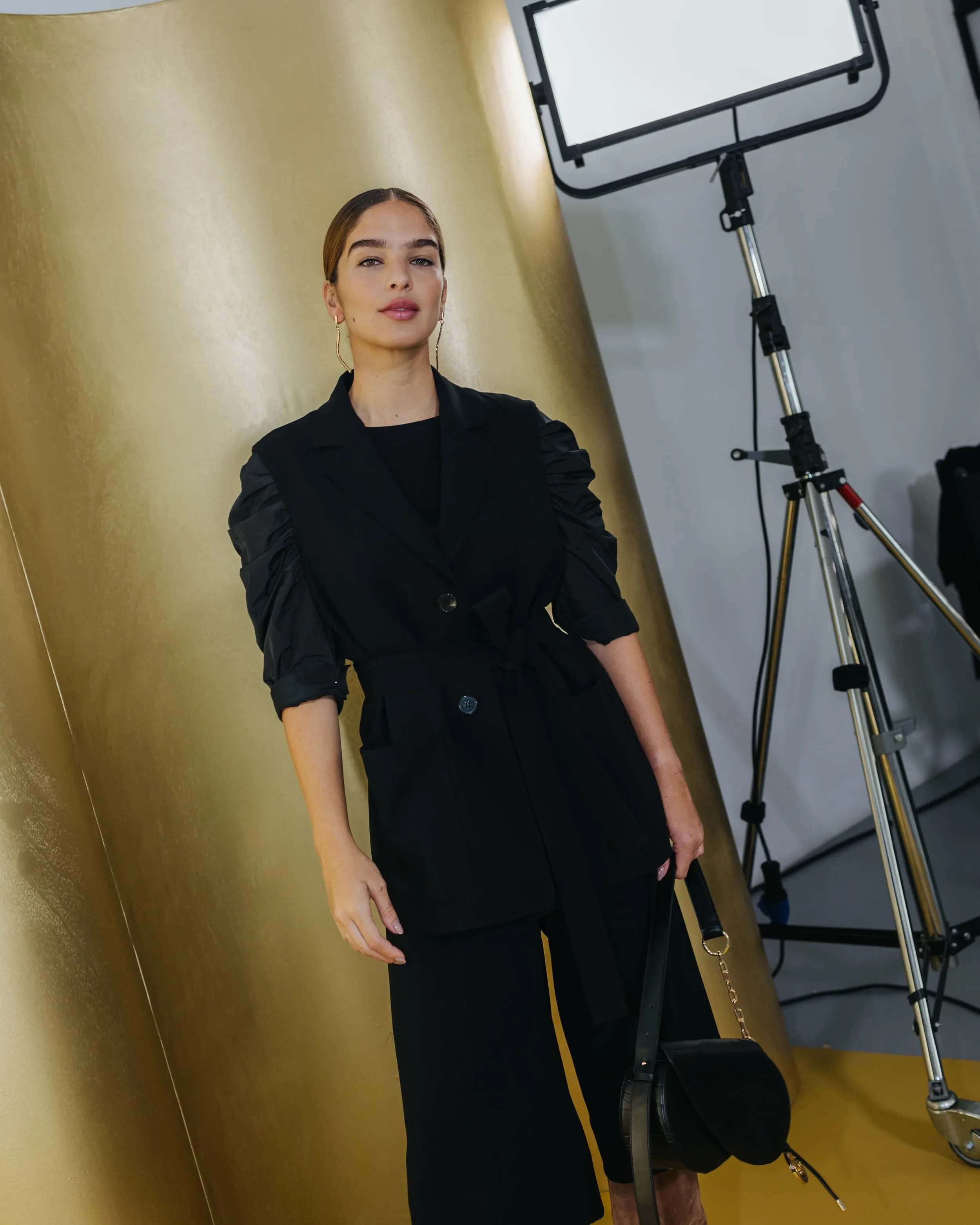 Modella nel backstage Marina Rinaldi indossa un completo sartoriale nero con gilet e maniche arricciate, posando davanti alla quinta dorata curva del set scenografico da studio.