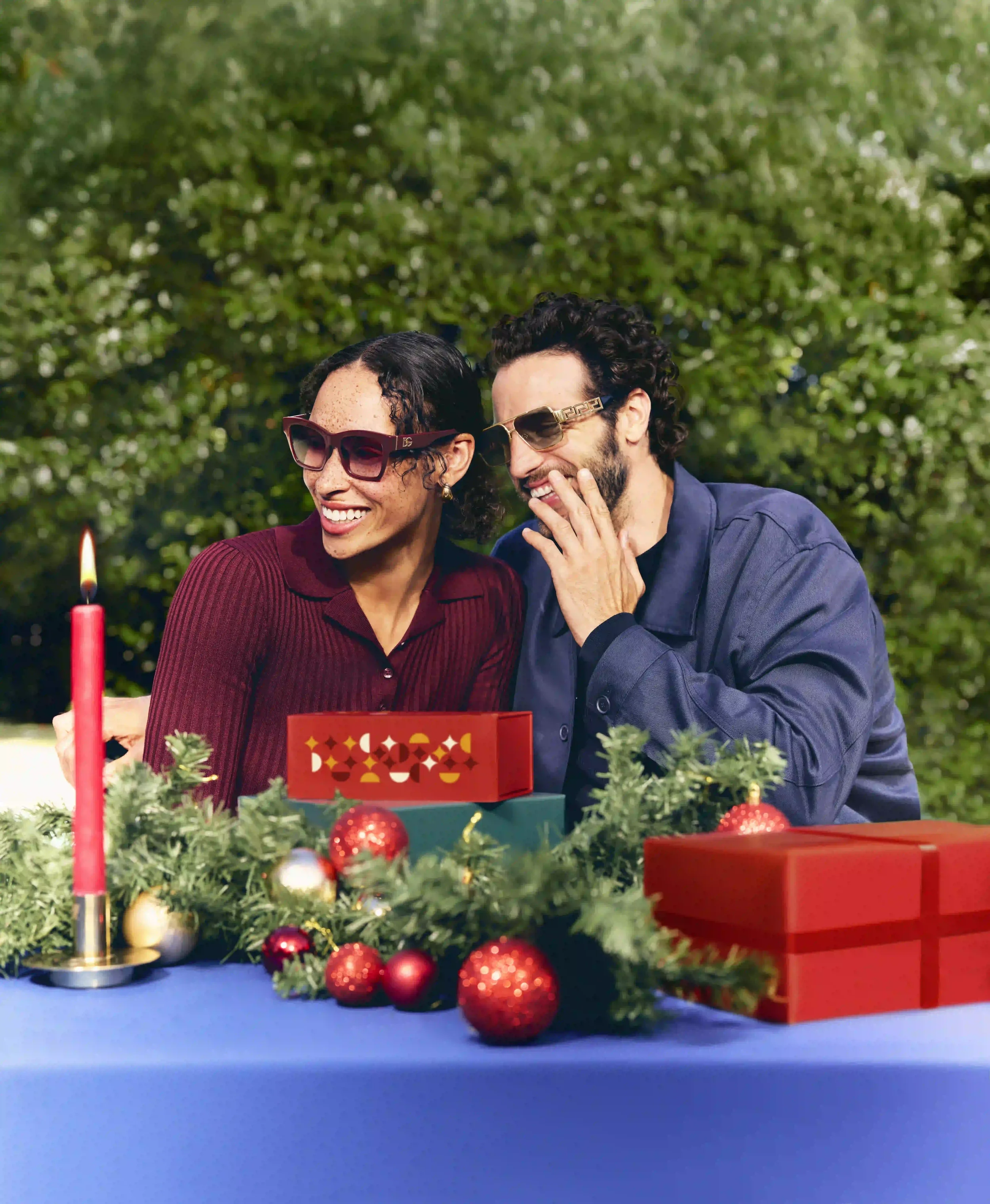 Scatto editoriale per la campagna di Natale Sunglass Hut con coppia al tavolo festivo decorato con candela rossa e ghirlande, mentre indossano occhiali da sole eleganti da uomo e donna.