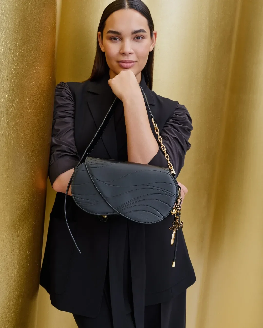 Primo piano di una borsa a spalla in pelle nera con impunture a onda e charm dorato MR, indossata da una modella davanti al pannello dorato curvo del set Marina Rinaldi.