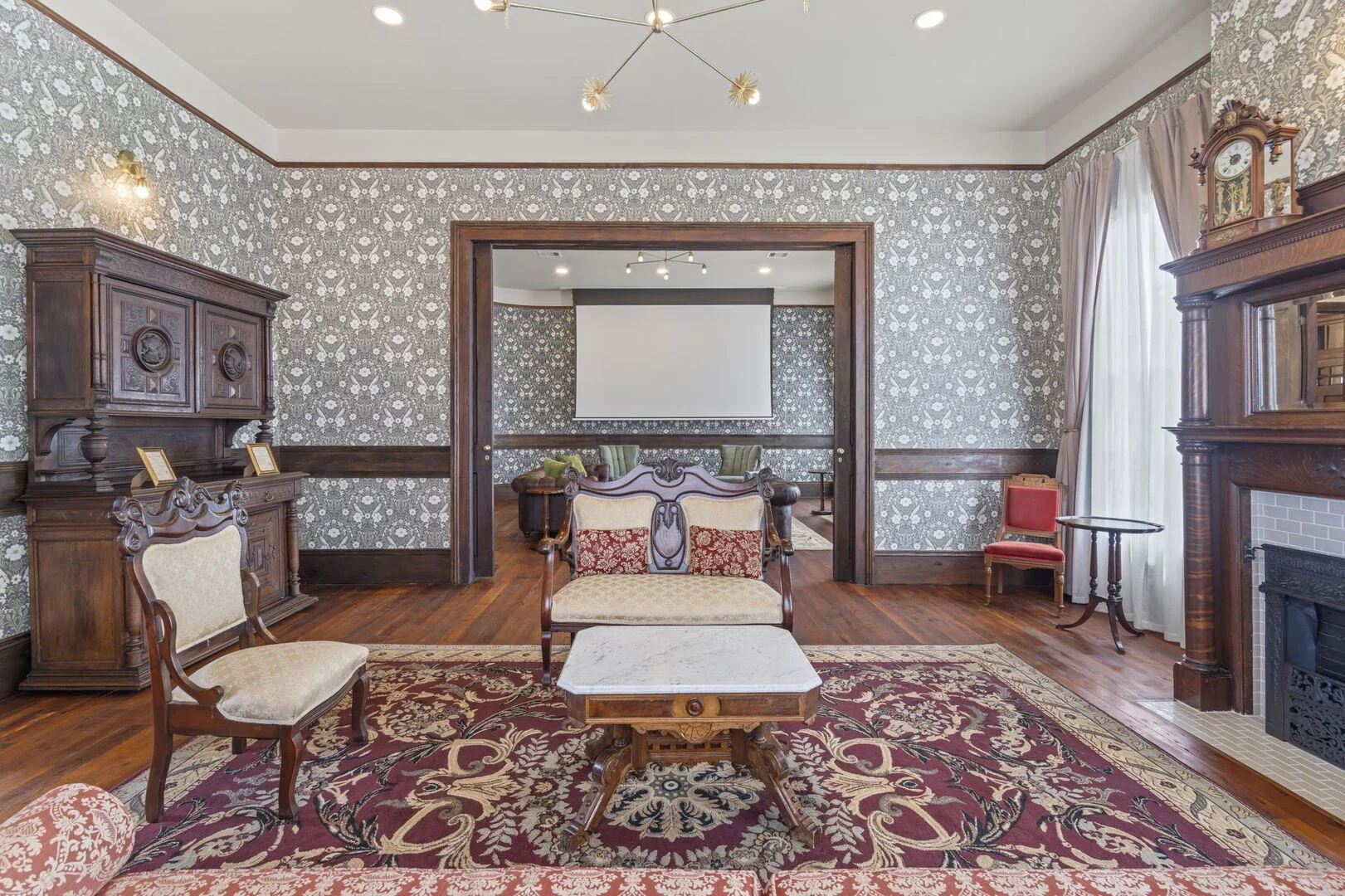 Restored Victorian Scarlet Parlor in the Mollie Walters House featuring hardwood floors, vintage furniture, a patterned rug, and fireplace. A doorway leads to the Daisy Parlor with a large projection screen and additional seating.