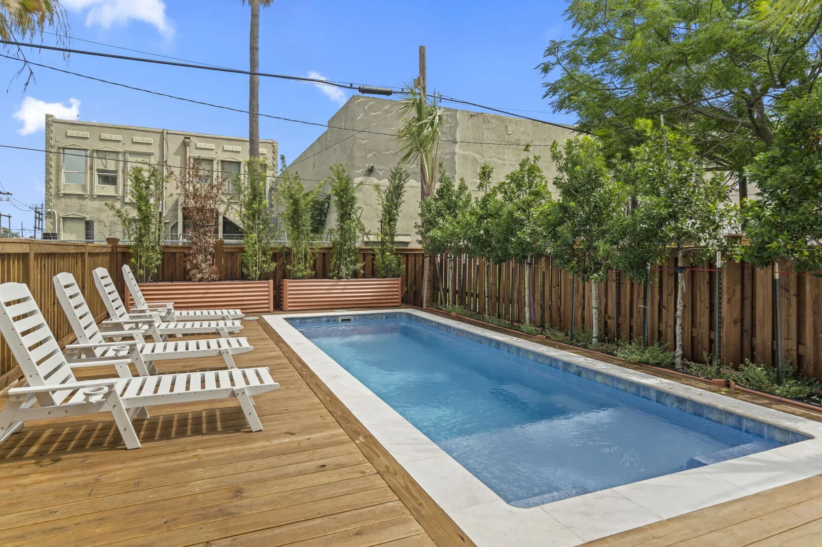 Mollie Walters House backyard with a rectangular swimming pool surrounded by a wooden deck, white lounge chairs, and green trees along a wooden fence. 