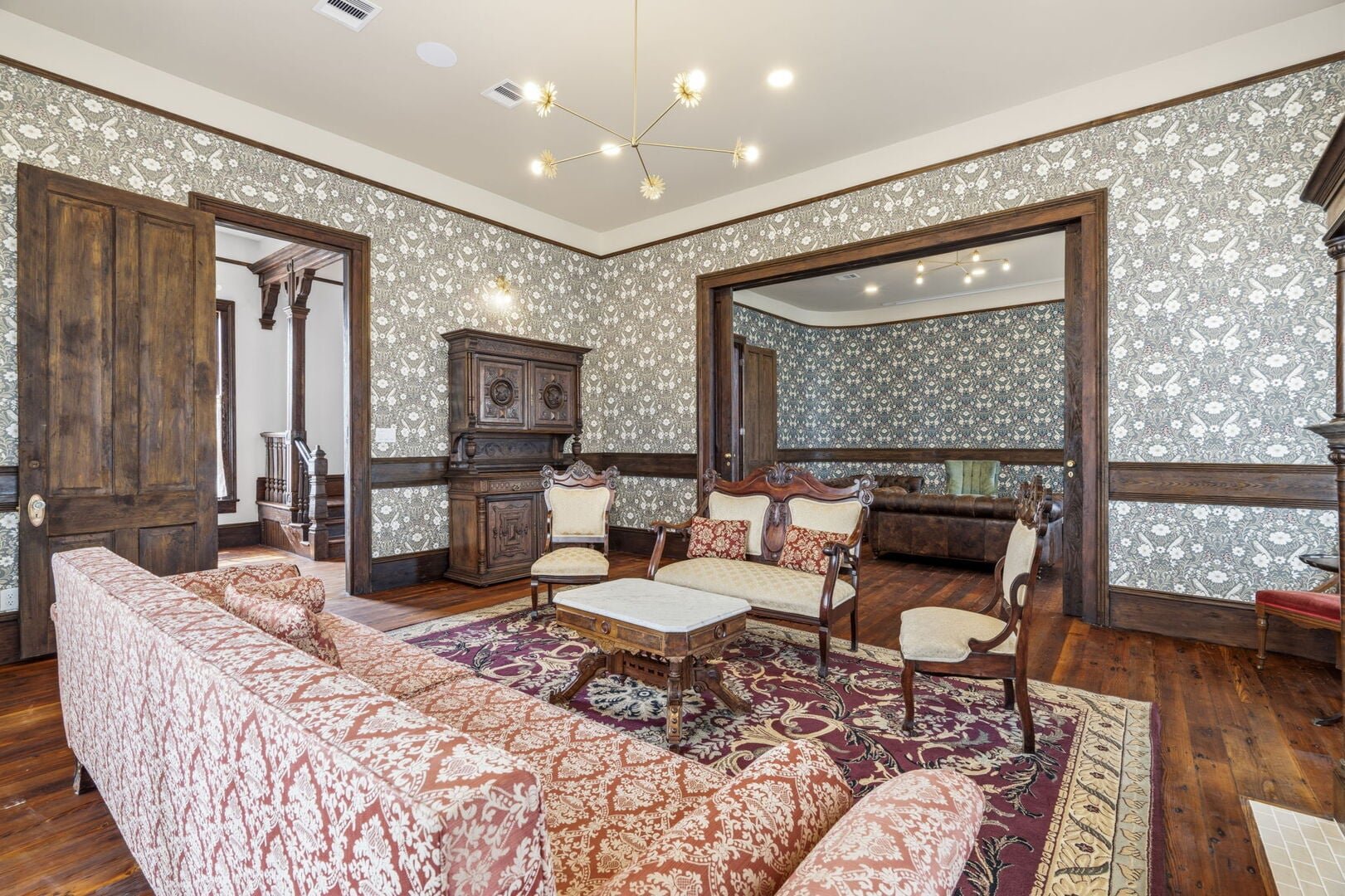 Restored Victorian Scarlet Parlor with floral wallpaper, wooden furniture including a sofa, chairs, and a cabinet, and area rugs. 