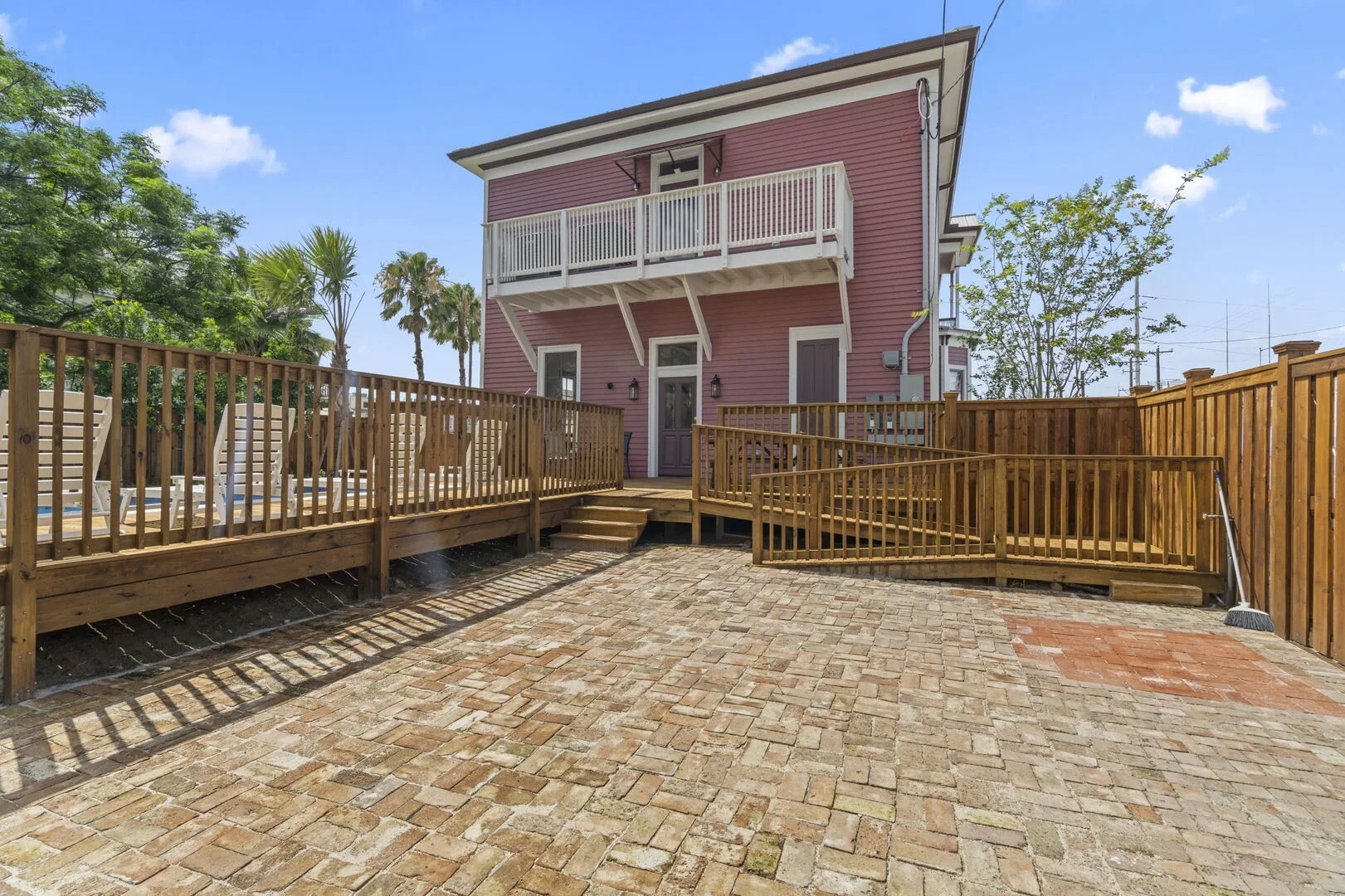 The restored 1886 Mollie Walters House backyard with brick patio, wooden deck with stairs, pink house with balcony, surrounded by wooden fence, with trees and blue sky in the background.