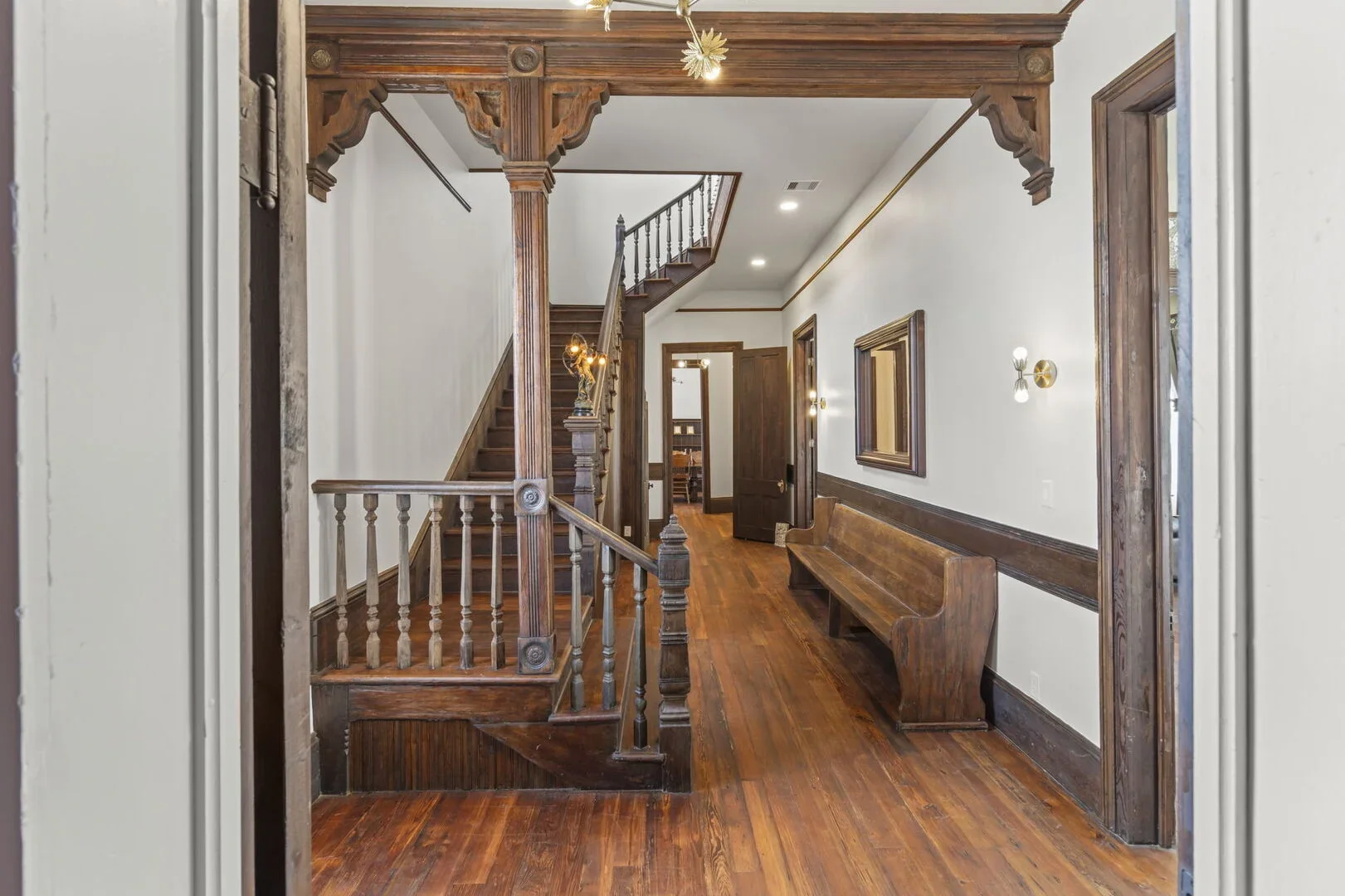 View of Mollie Walters House Victorian interior hallway with wooden staircase, wooden bench, and decorative light fixtures, and hardwood flooring.
