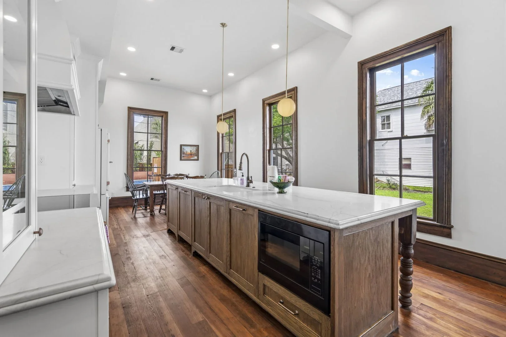 Mollie Walters House Kitchen with wooden cabinets, white marble island countertop with sink, black microwave, large windows with wooden trim, hardwood floor, and dining area in the background.