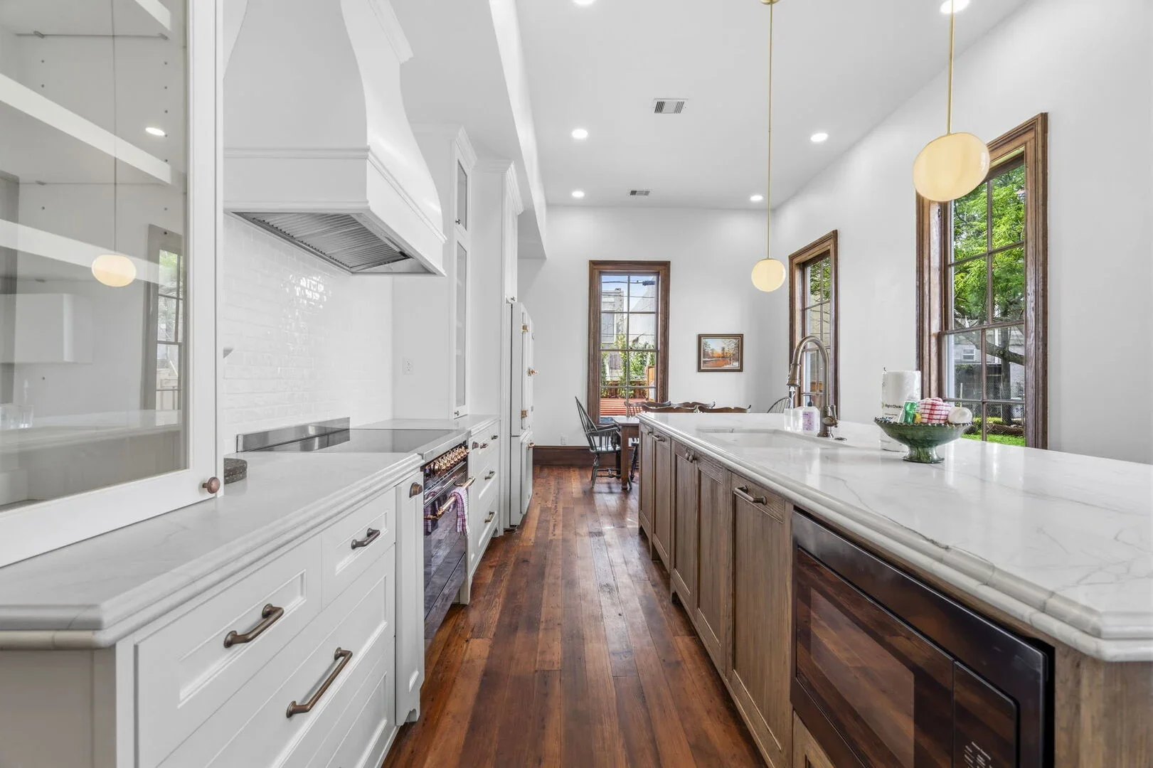 Mollie Walters House Kitchen with white cabinets, a large island with a marble countertop, and wooden floors pendant lights hanging over the island, and a dining table with chairs