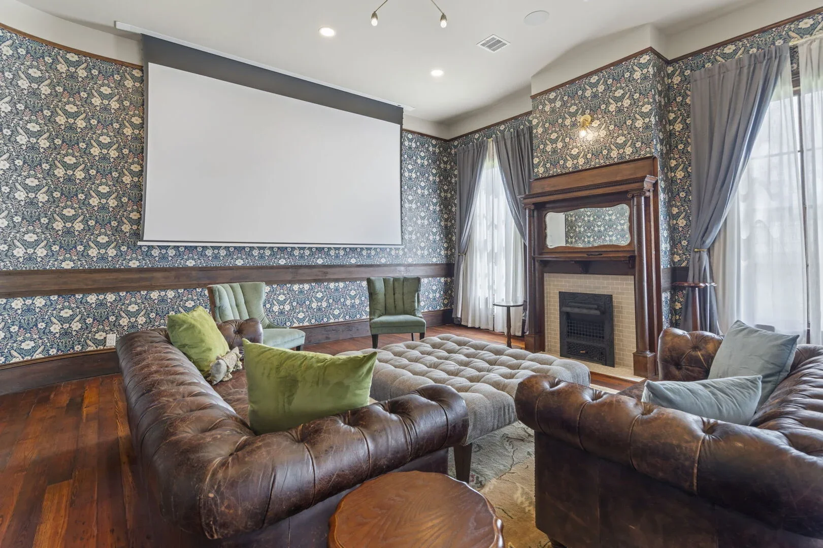 Mollie Walters Daisy Parlor with floral wallpaper, leather sofa, tufted ottoman, two green chairs, fireplace, and a large retractable projector screen.