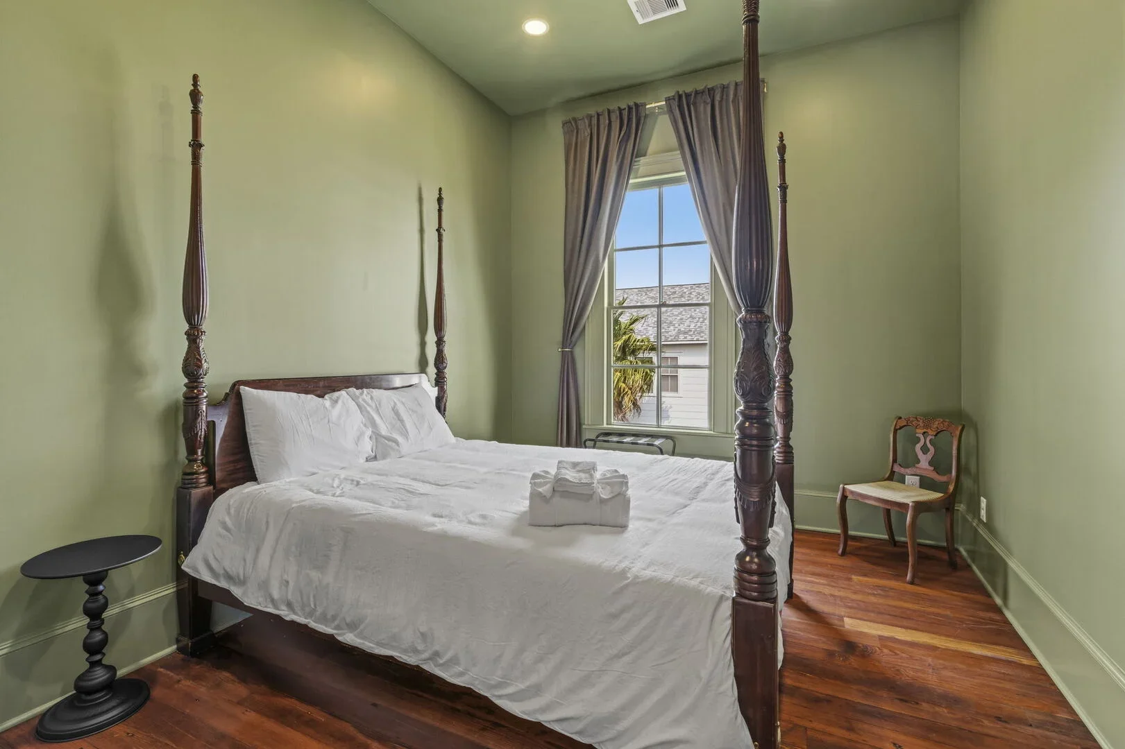 The Mollie Walters Olive Chamber with a wooden four-poster bed queen size bed, a wooden chair in the corner, and a window with view of a palm tree.