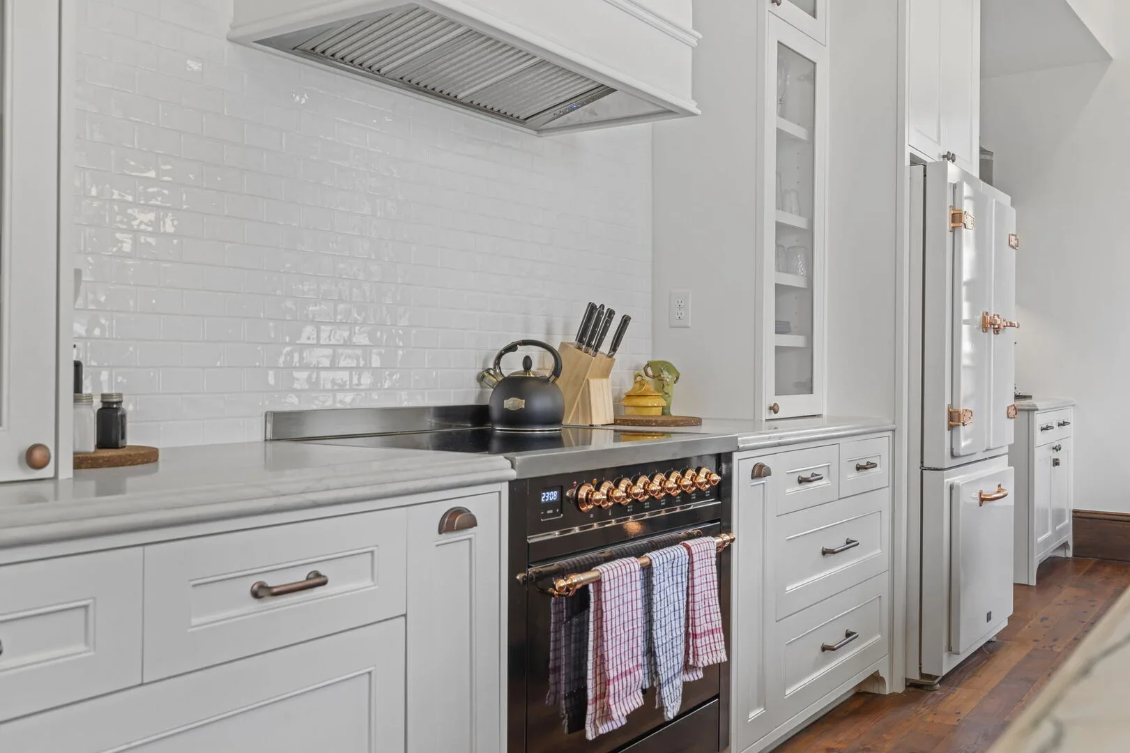 Mollie Walters House Kitchen with copper accents, stove with kitchen towels, black kettle, knife block, and white cabinetry.