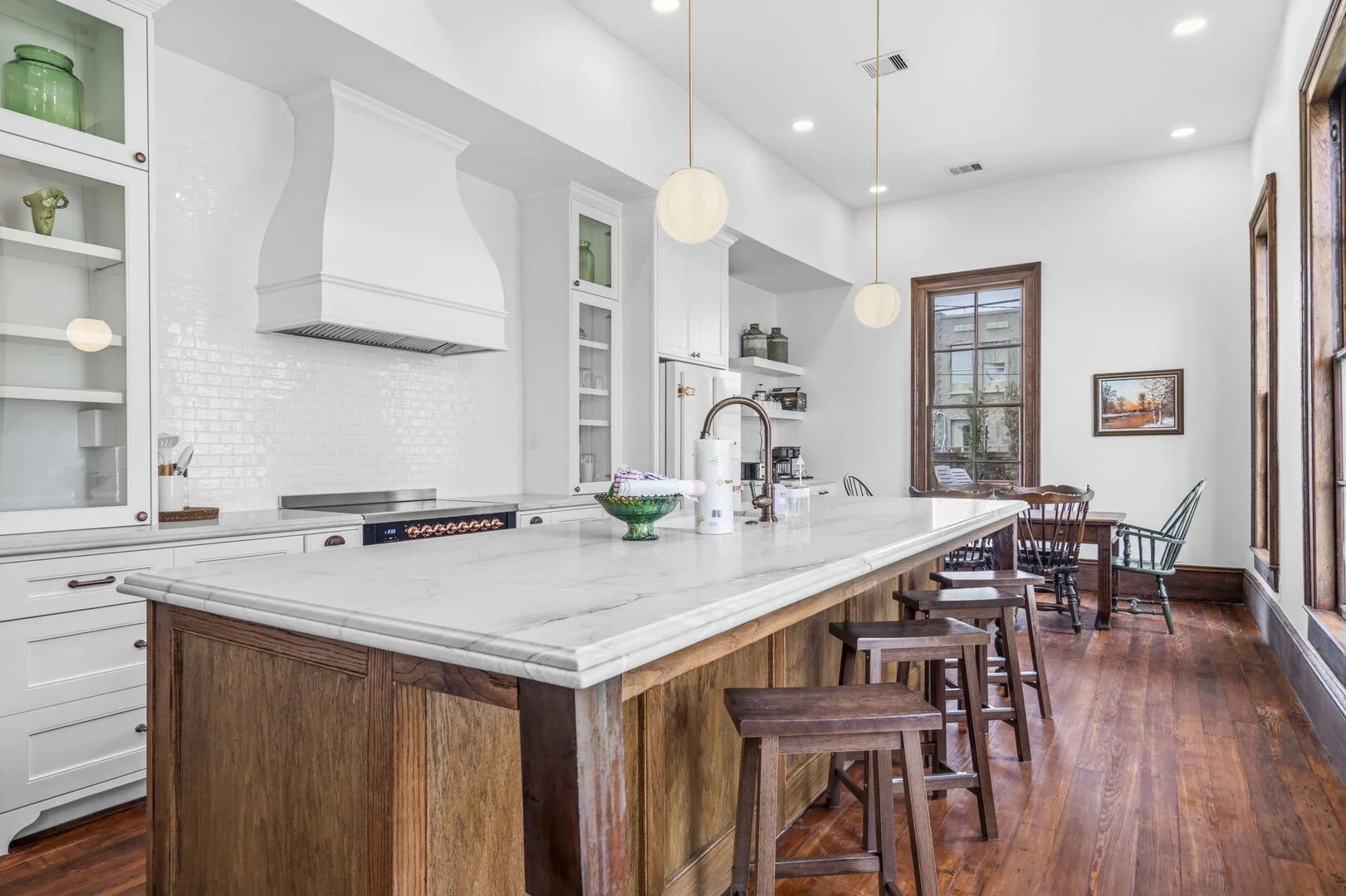 Mollie Walters House Kitchen with white cabinets, a white marble island, wooden stools, and four round pendant lights hanging from the ceiling hardwood flooring, and a dining table with chairs in the corner.