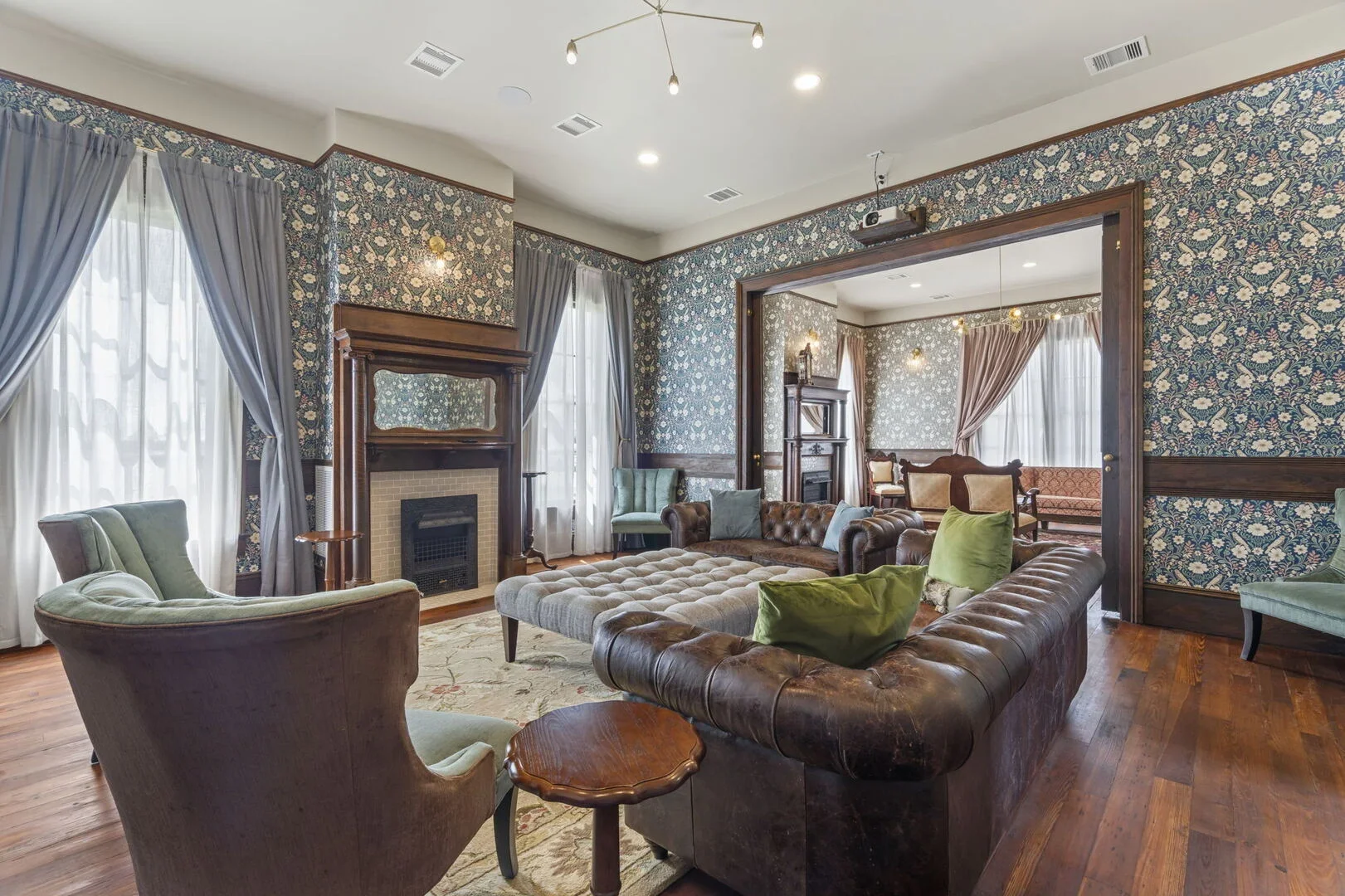 Mollie Walters Daisy Parlor with floral wallpaper, leather sofa, tufted ottoman, two green chairs, and fireplace