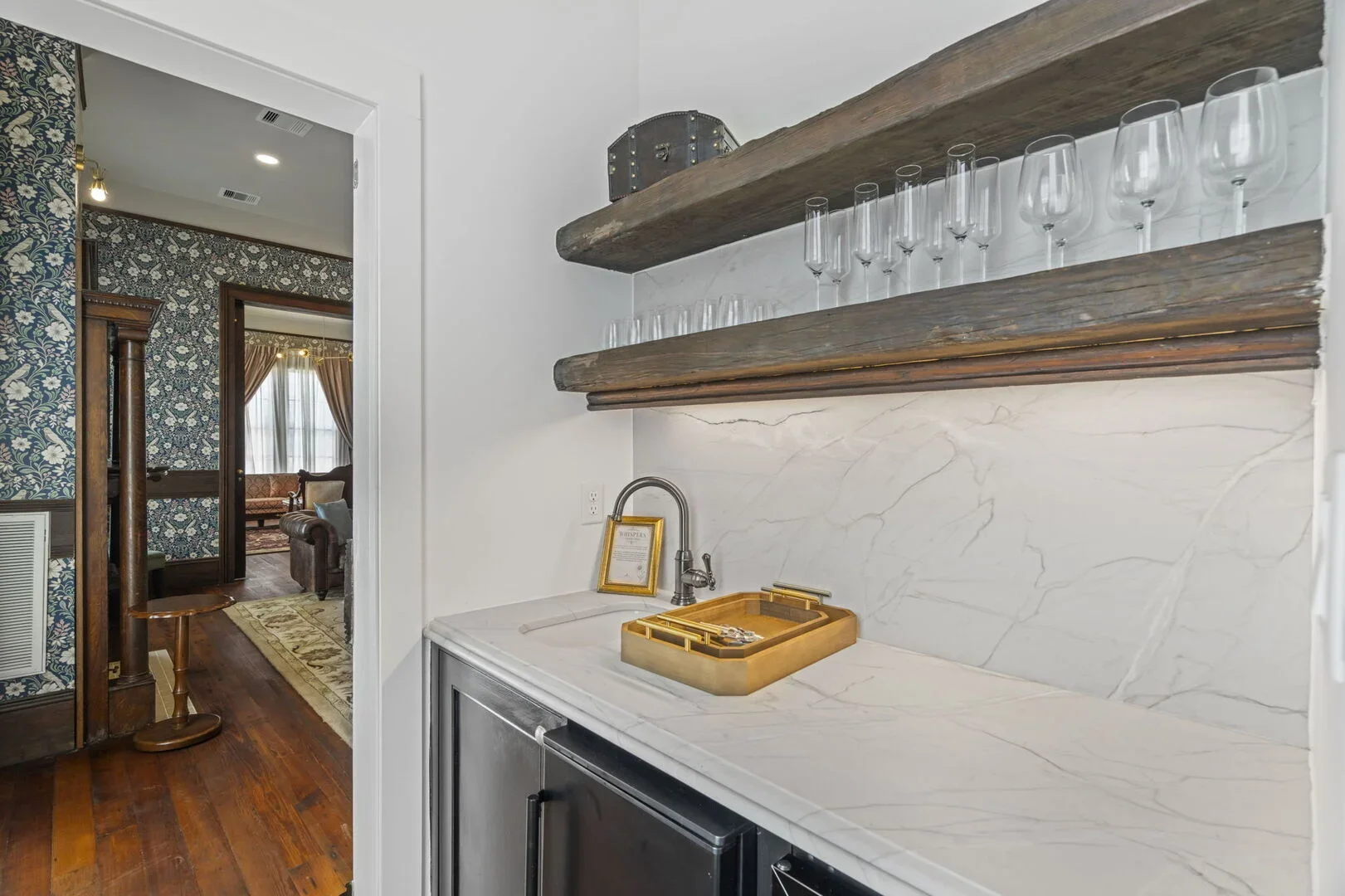 Mollie Walters House Kitchen bar leading to the Daisy Parlor with two rustic wooden shelves holding glassware, marble countertop, and sink