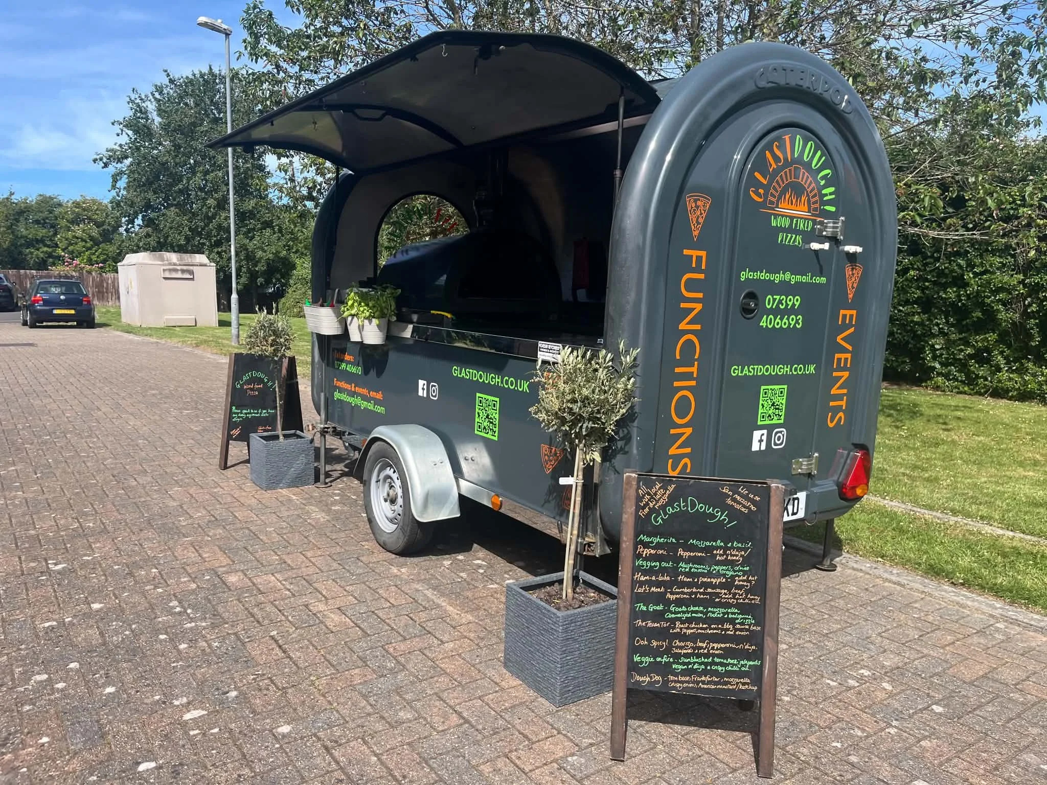 A mobile food trailer with colorful menu signs and a chalkboard menu, parked on a paved surface with trees and cars in the background, selling wood-fired pizza.