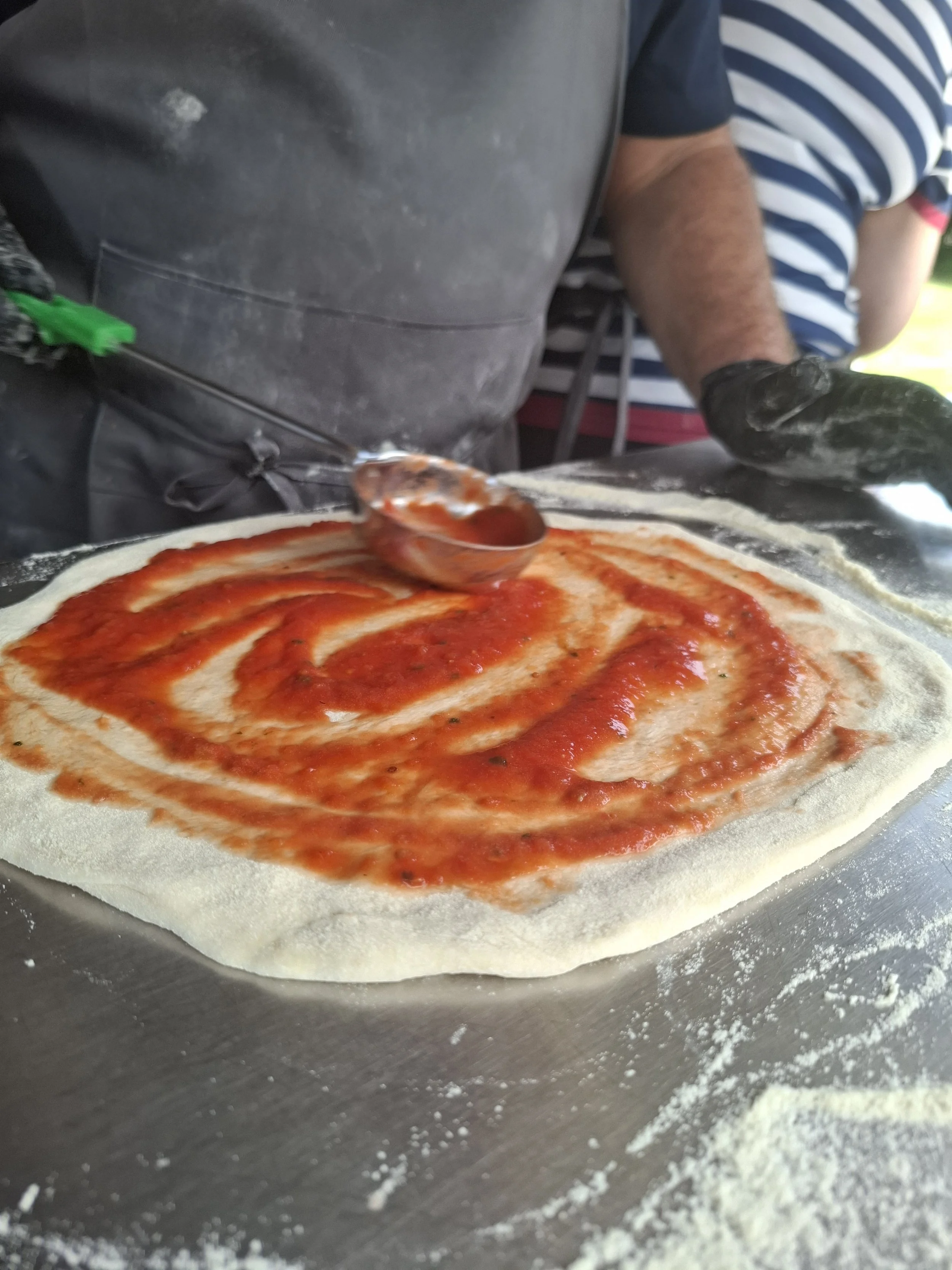 Close-up of a pizza dough with tomato sauce topped on it, on a floured surface, with a person in striped shirt preparing to make pizza.