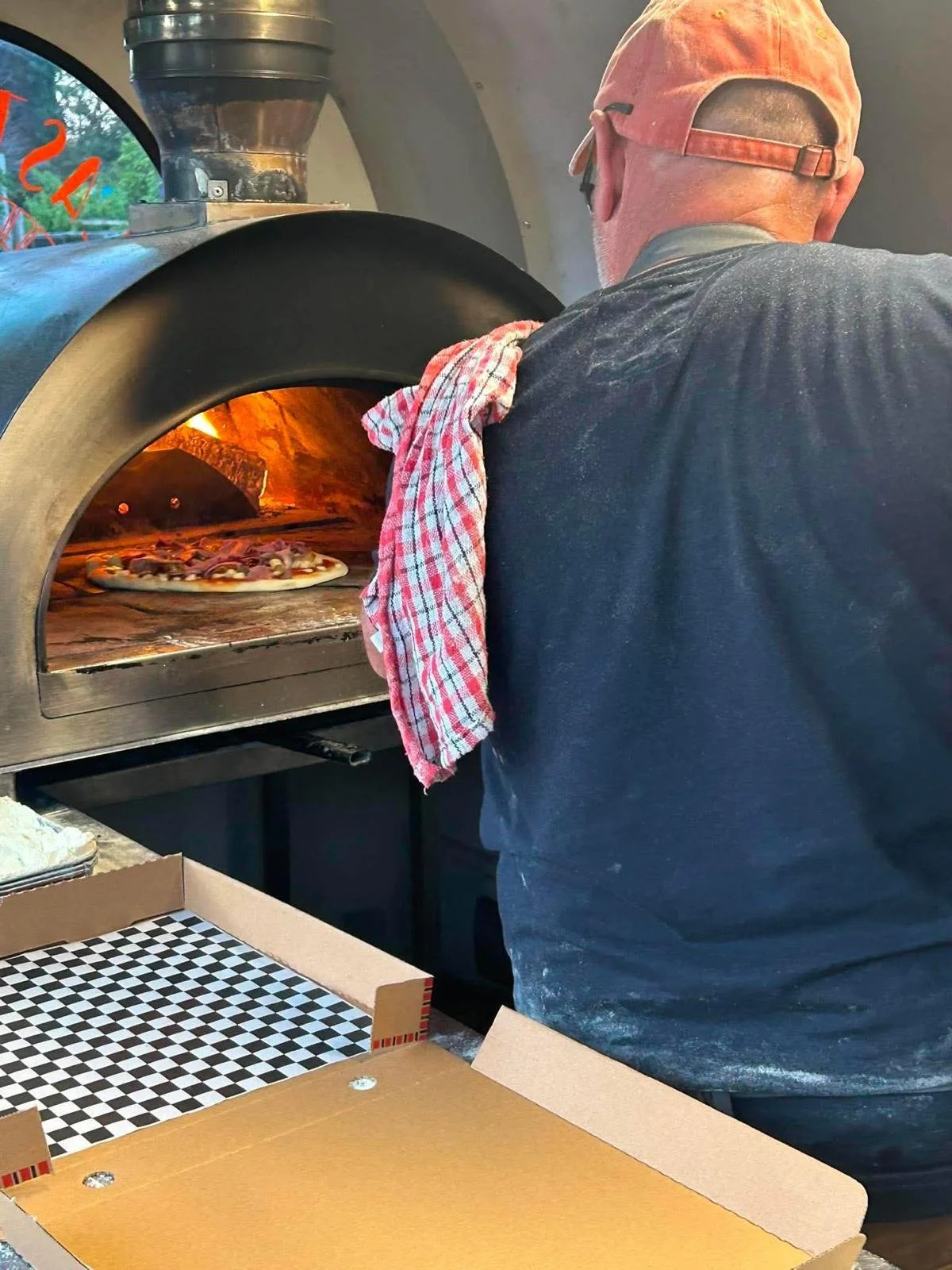 A person wearing a pink cap and black shirt is removing a pizza from a stone oven, with a red checkered towel hanging on their shoulder.