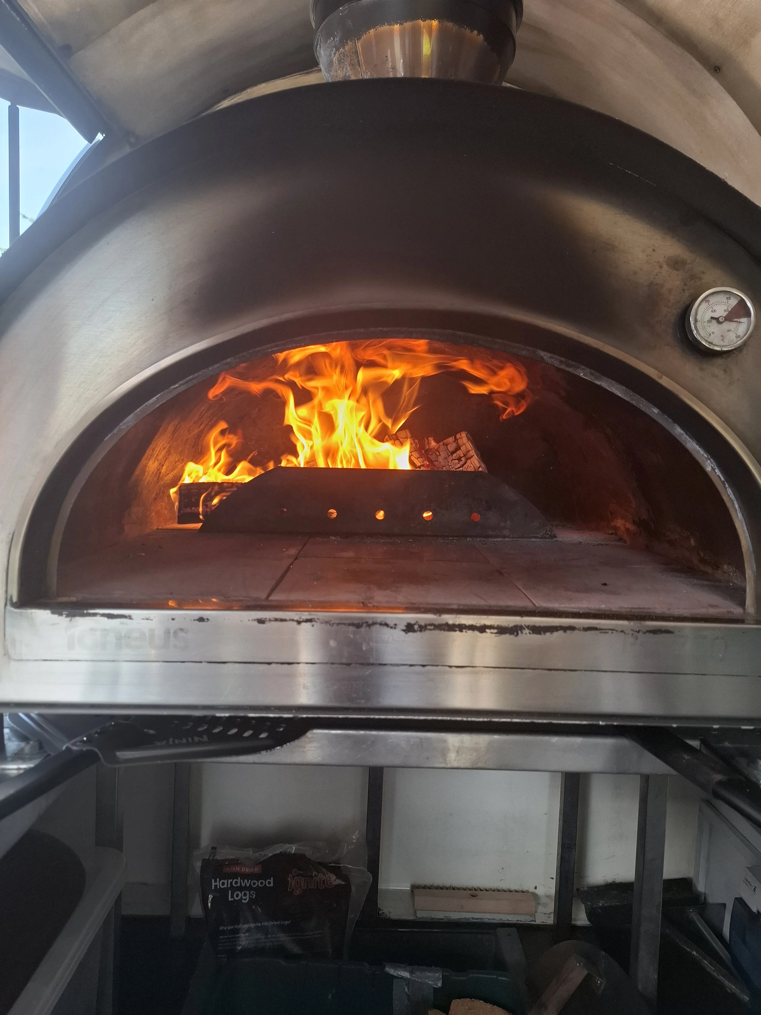 A commercial pizza oven with flames inside and a thermometer on the front, used for baking pizzas.