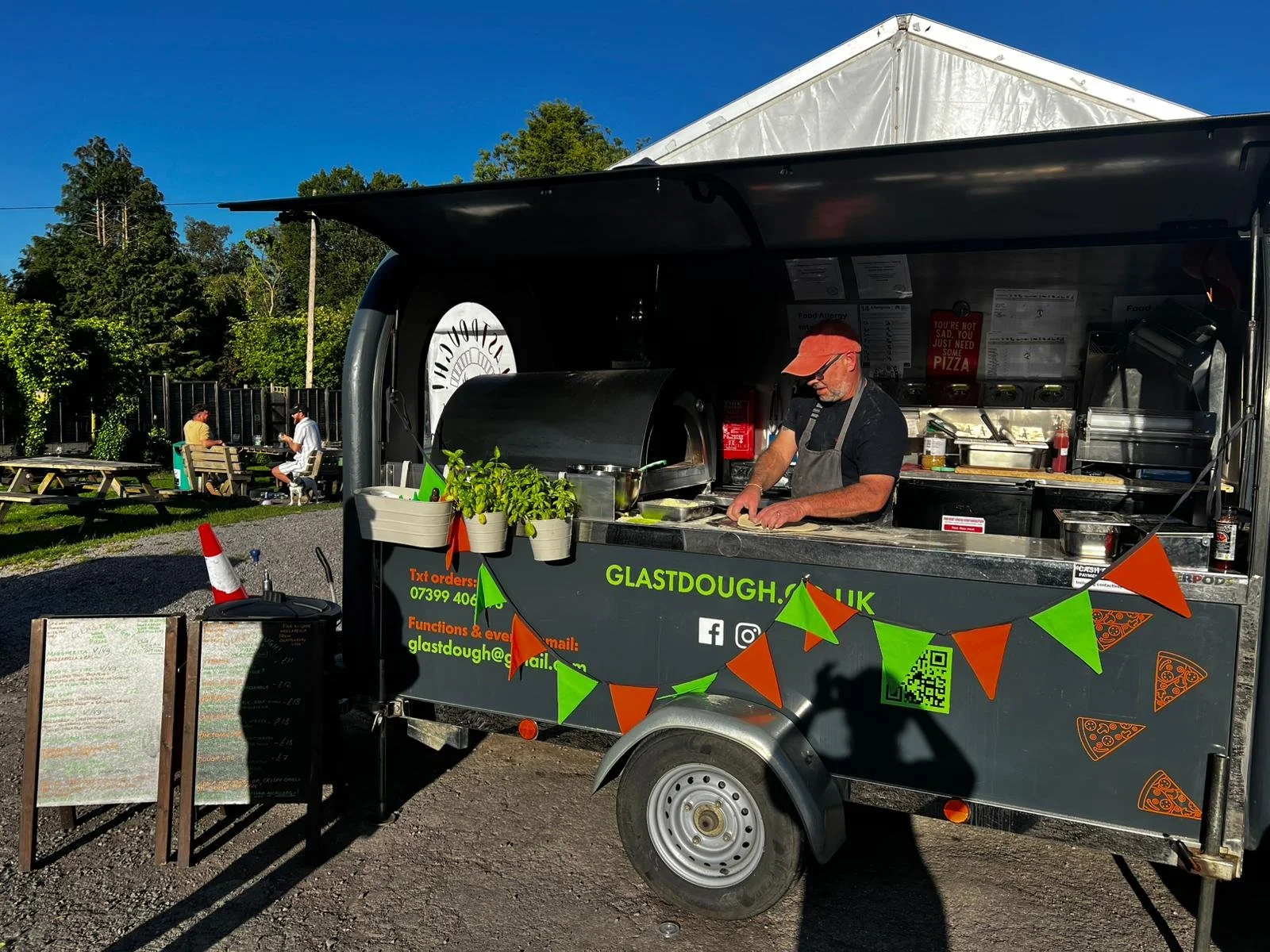 A mobile pizza food truck with a man preparing pizza on a counter outside, decorated with green and orange bunting, in a park setting with trees and people sitting at picnic tables in the background on a sunny day.